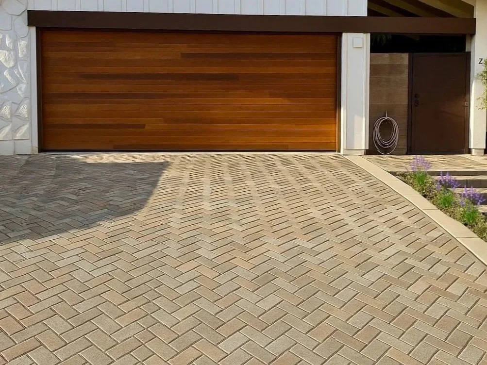 Concrete driveway with a herringbone pattern leading to a brown garage door, with a side garden and hose reel near the doorway.