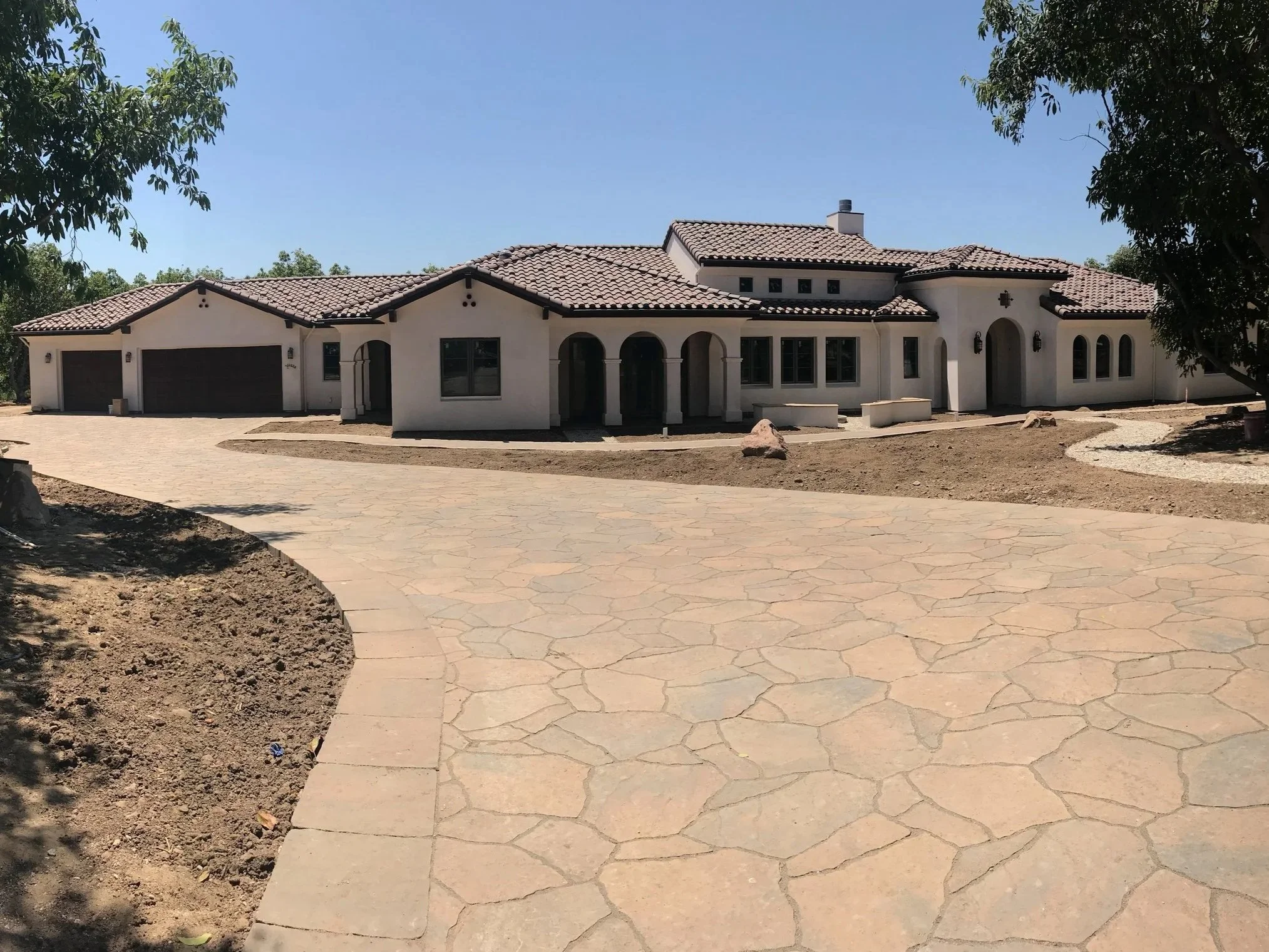 A large, white stucco house with a tiled roof, arched windows, and multiple garage doors. The house is surrounded by a paved driveway and some trees.
