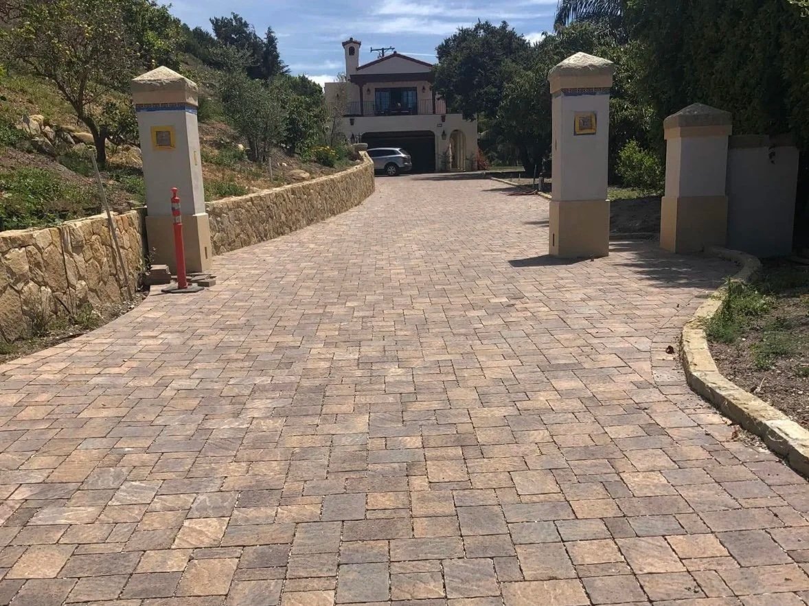 A paved driveway leading to a house, flanked by stone wall on the left and a curved curb on the right, with trees and greenery around.
