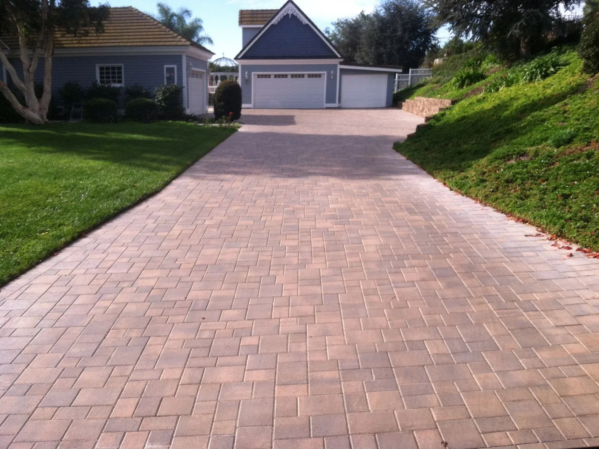 A driveway made of rectangular paving stones leads to a garage with three doors. On either side of the driveway, there are well-maintained green lawns and shrubbery. In the background, two houses with blue siding and brown shingle roofs, and trees ar