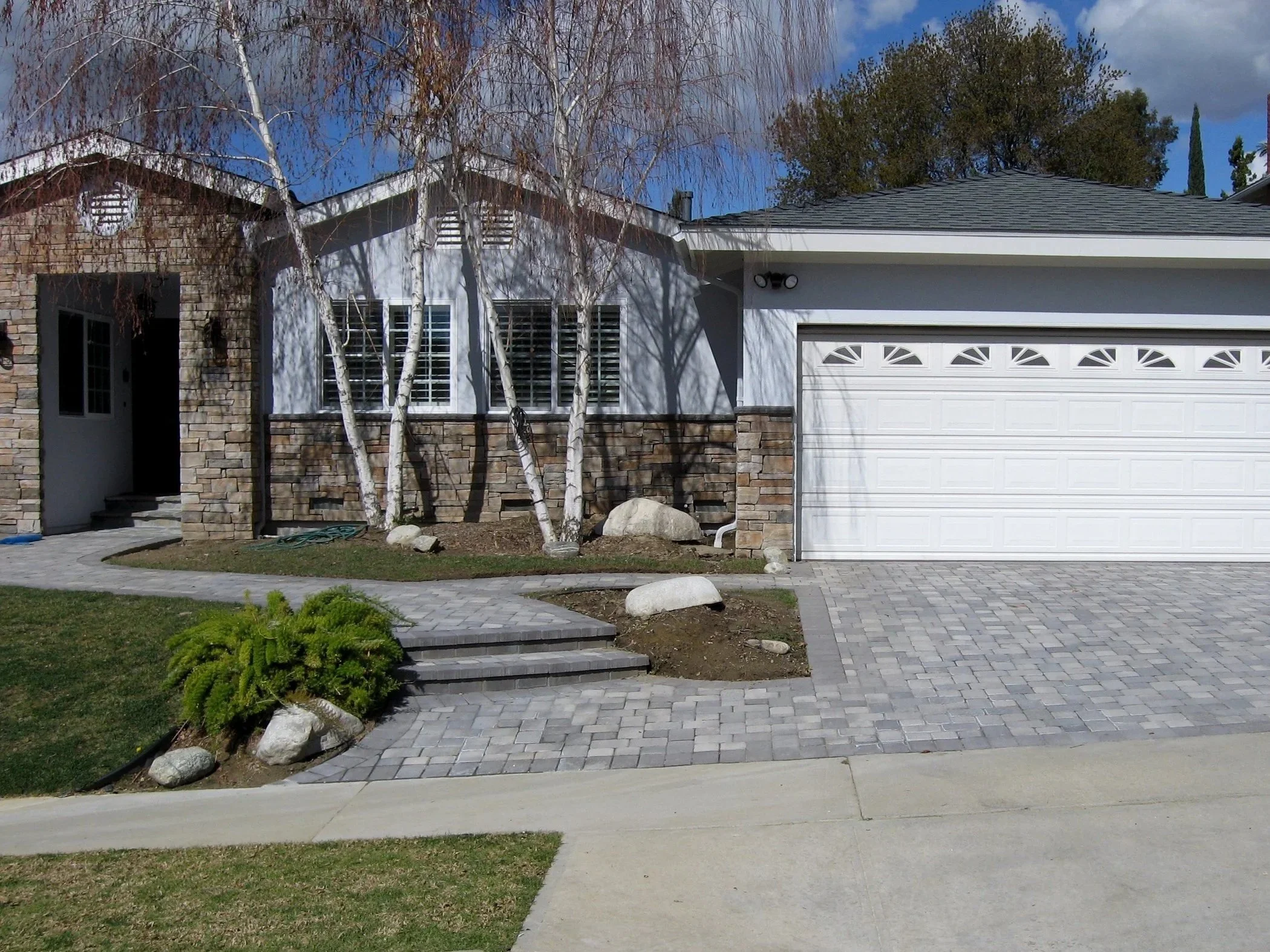 Exterior view of a house showing a stone and white siding facade, front yard with trees, paved walkway, and a garage door.