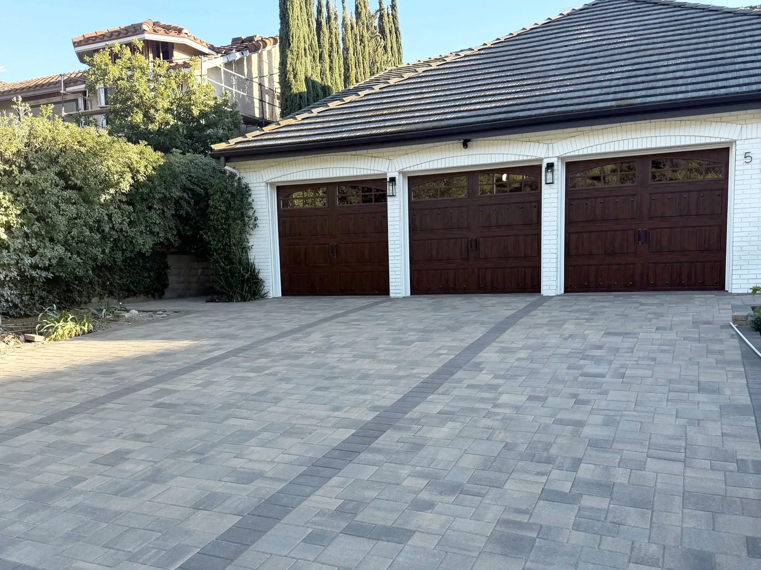 A residential driveway with brick pavers in a pattern, leading to a white house with dark wooden garage doors, surrounded by greenery and trees.