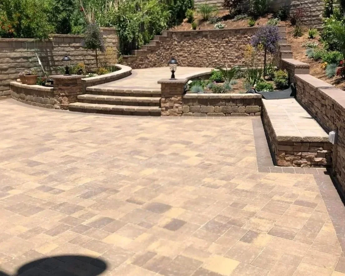 A multi-level outdoor patio area with beige stone pavers, stone steps, and brick retaining walls. There are small plants and shrubs along the walls and decorative lights.