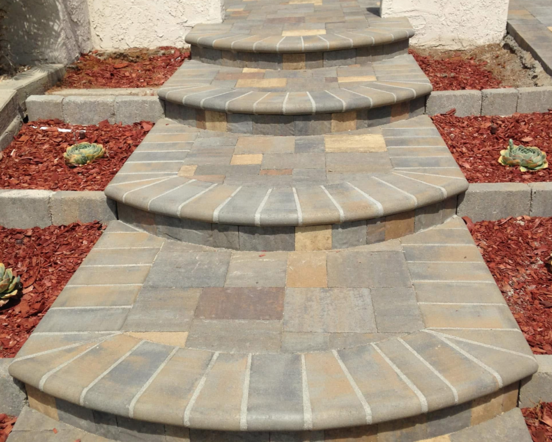 Stone outdoor stairs with curved risers and brick pattern steps, flanked by red mulch and small succulents.