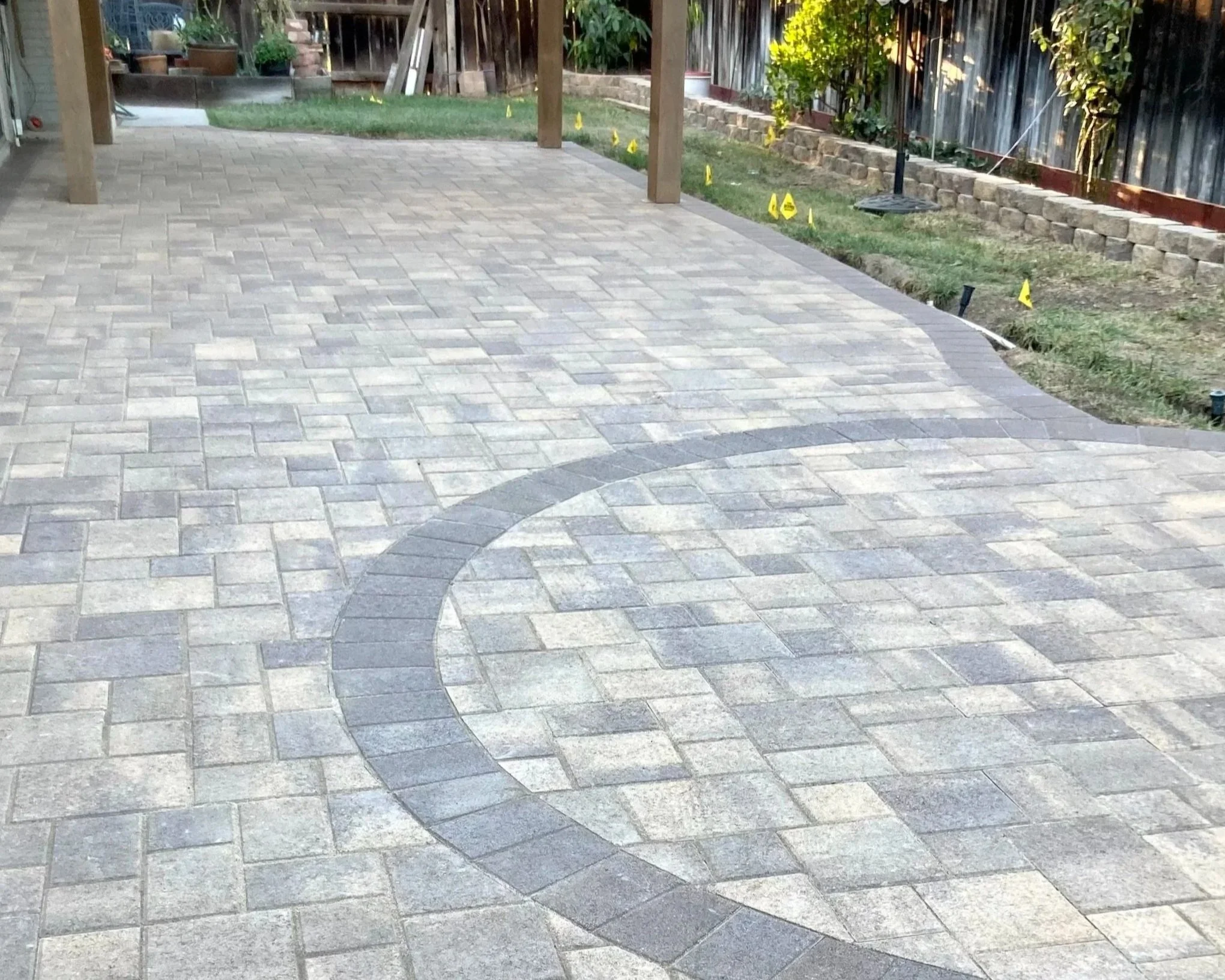 Backyard patio with newly installed stone pavers, surrounded by a garden bed with yellow flags marking underground utilities, a raised brick border, and a wooden pergola.