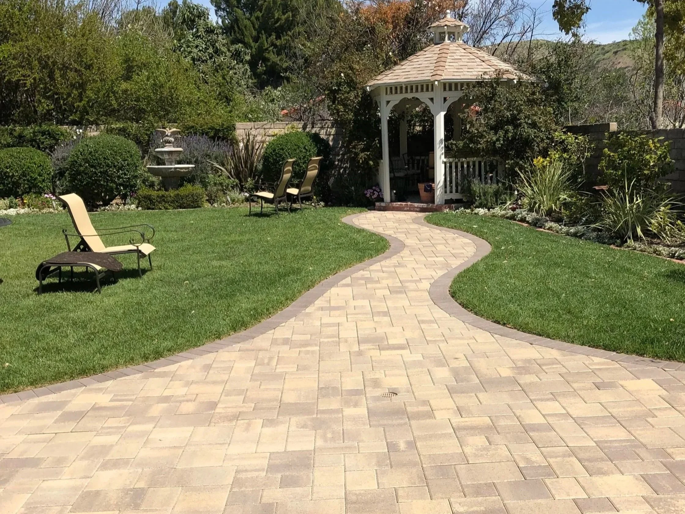 A backyard garden with a paved winding walkway, green lawn, chairs, a gazebo, a fountain, and various trees and shrubs.