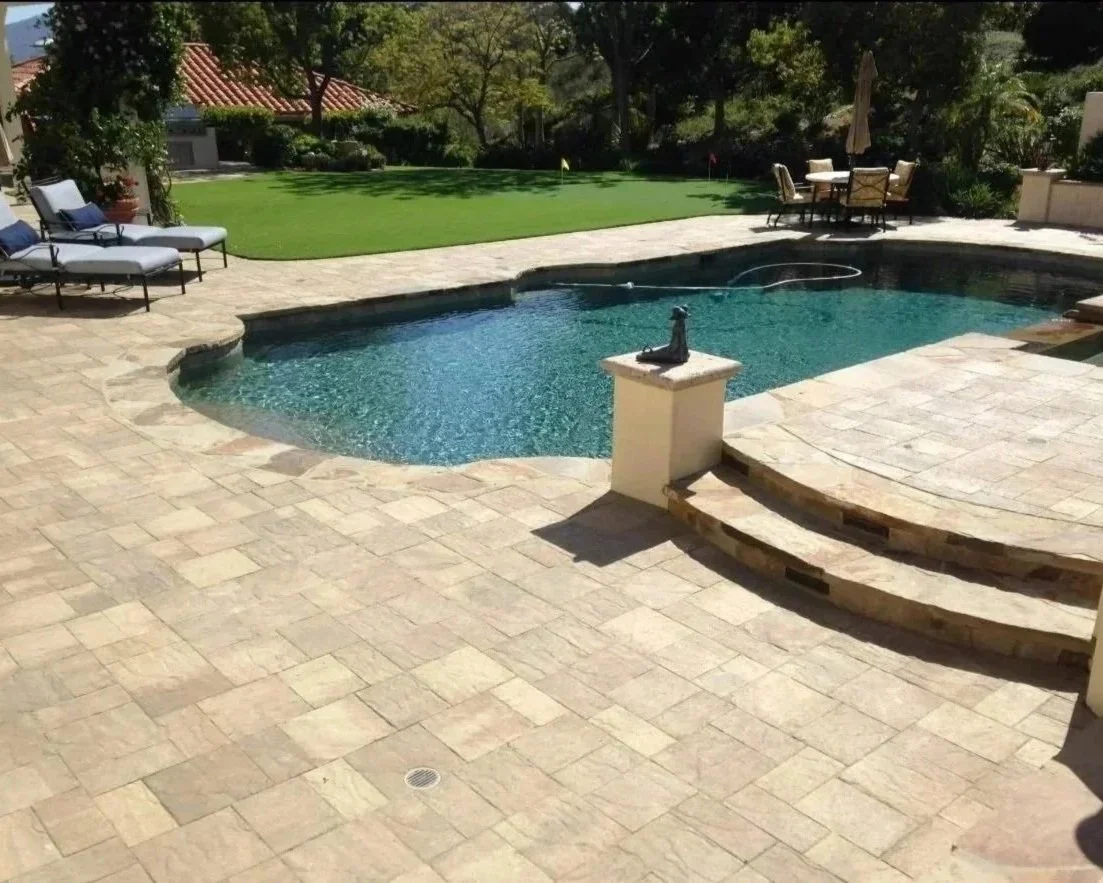 Backyard with swimming pool, lounge chairs, a round table with chairs and umbrella, green lawn, and trees.