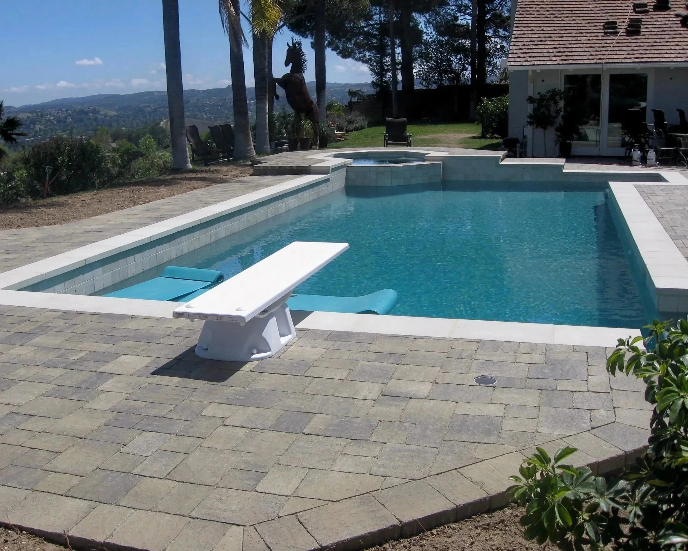 A swimming pool with a white edge and a built-in spa is in the backyard of a house. The pool area has a paver stone deck, and a lounge chair with a blue cushion. In the background, there are trees, including tall palm trees, and a view of hills under
