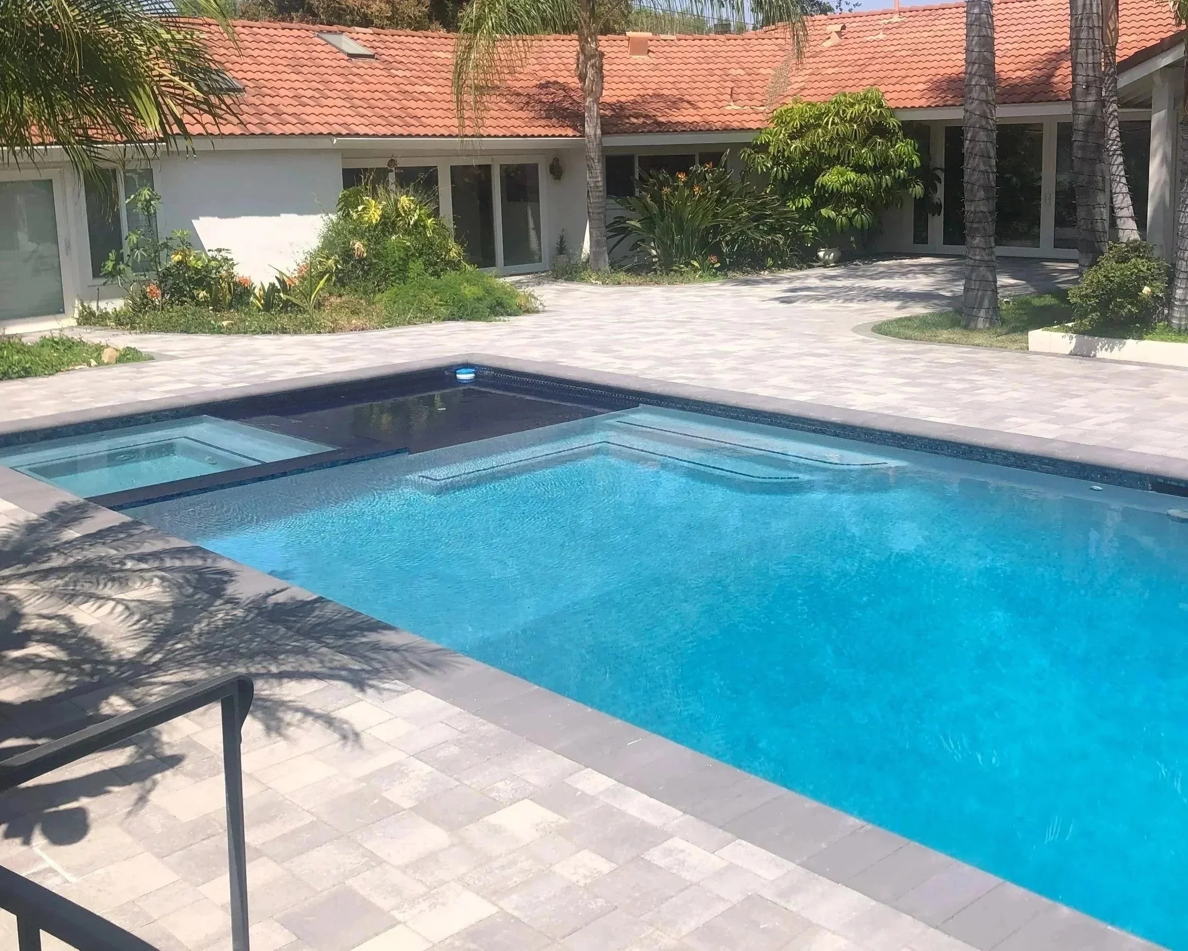 A backyard scene featuring a swimming pool with a clear cover, surrounded by gray stone paving. There are sun loungers nearby, some shadowed by trees. In the background, there is a house with a red-tiled roof, surrounded by lush green plants and palm