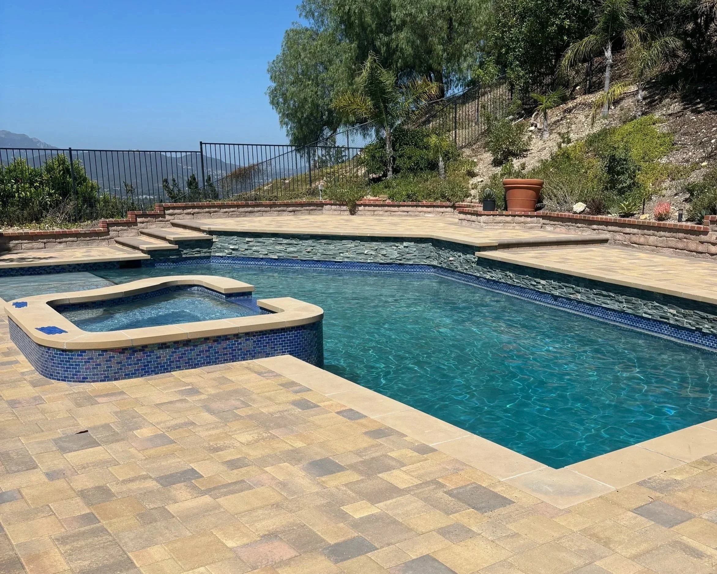 A backyard with a swimming pool, hot tub, stone patio, brick wall, potted plants, and a hillside with trees and a metal fence in the background.