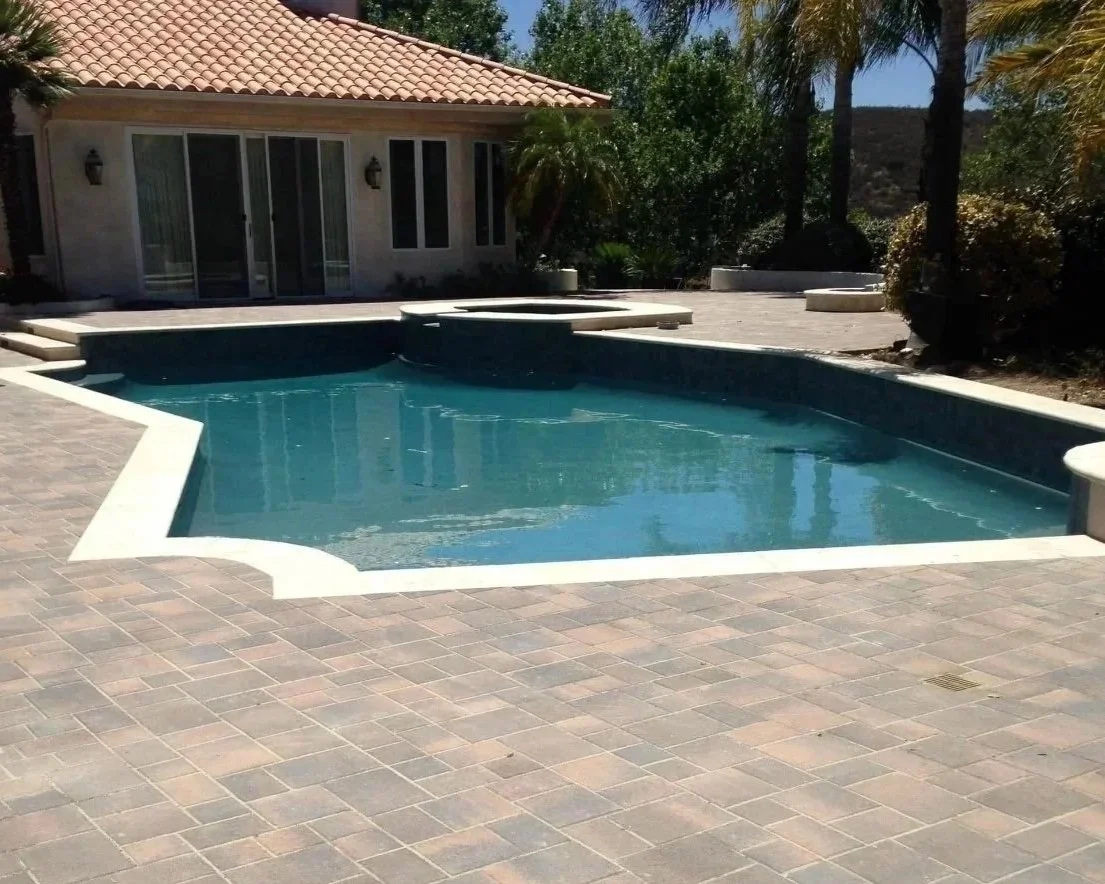 Residential backyard with a swimming pool and hot tub, paved patio, house with sliding glass doors, and surrounding trees and plants.