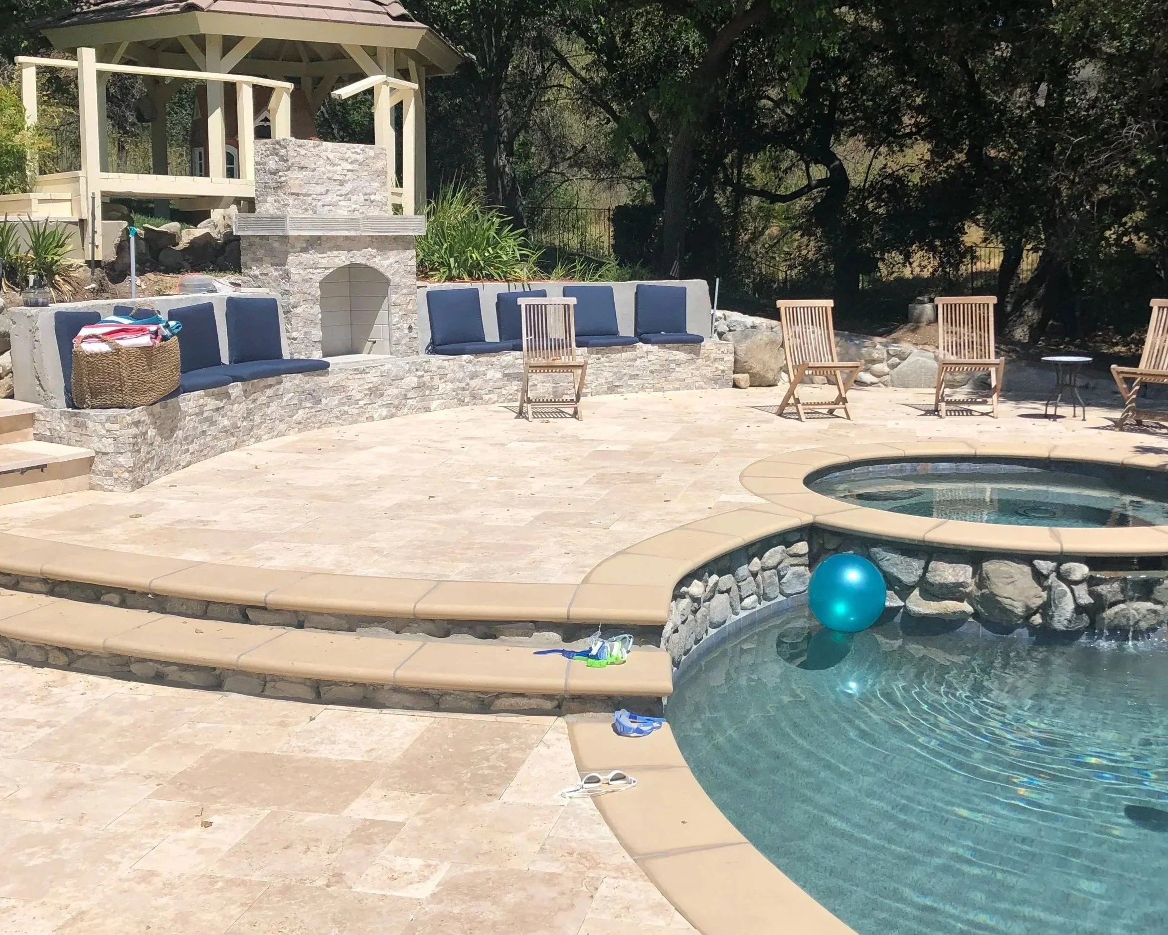 Backyard pool area with a swim-up bar, blue cushioned chairs, wooden chairs, and a small round table, surrounded by trees and a relaxed outdoor setting.