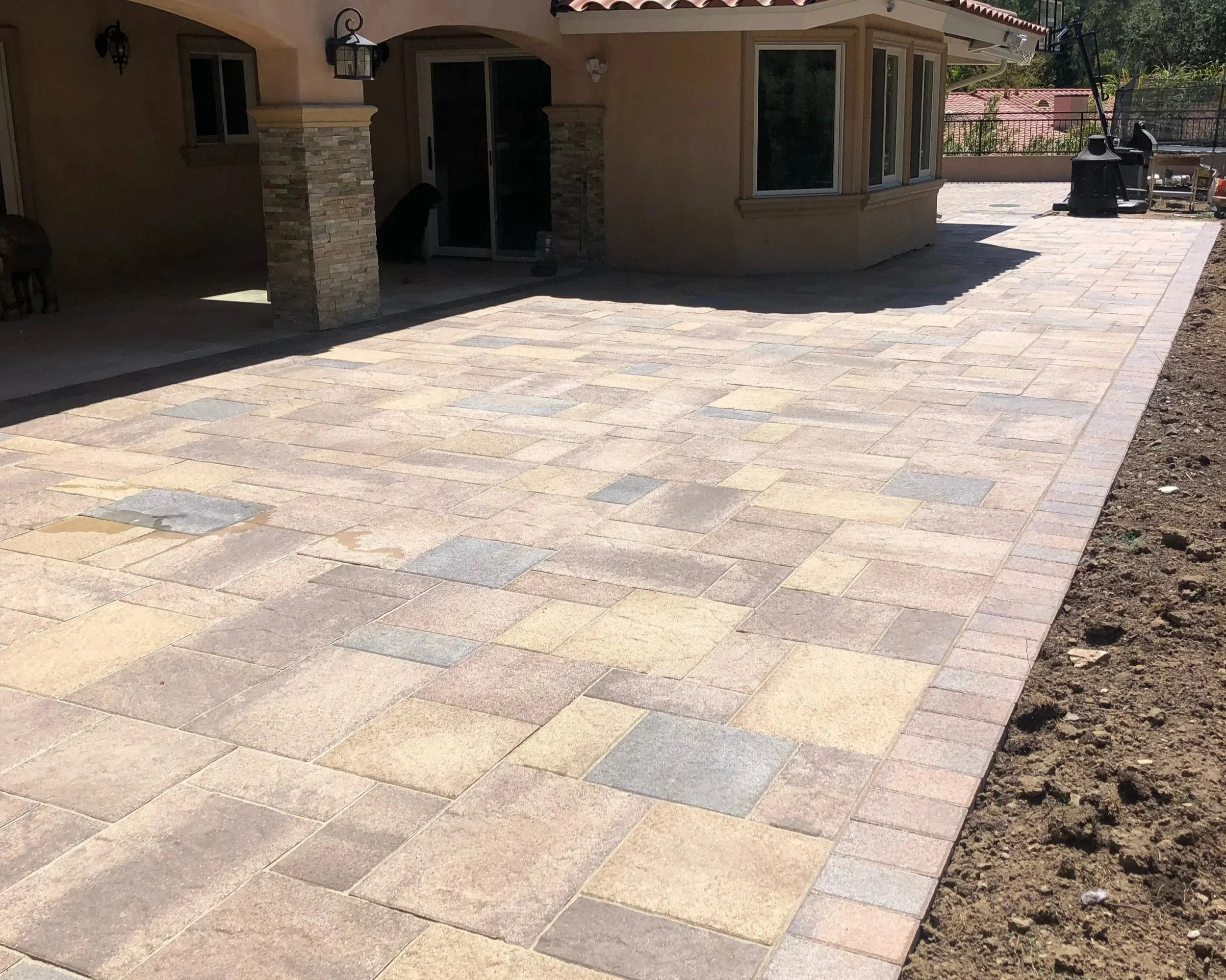 A newly paved backyard with interlocking stone patio, house with beige stucco walls, large windows, and a covered patio area with stone pillars. A grill and outdoor furniture are visible in the background.