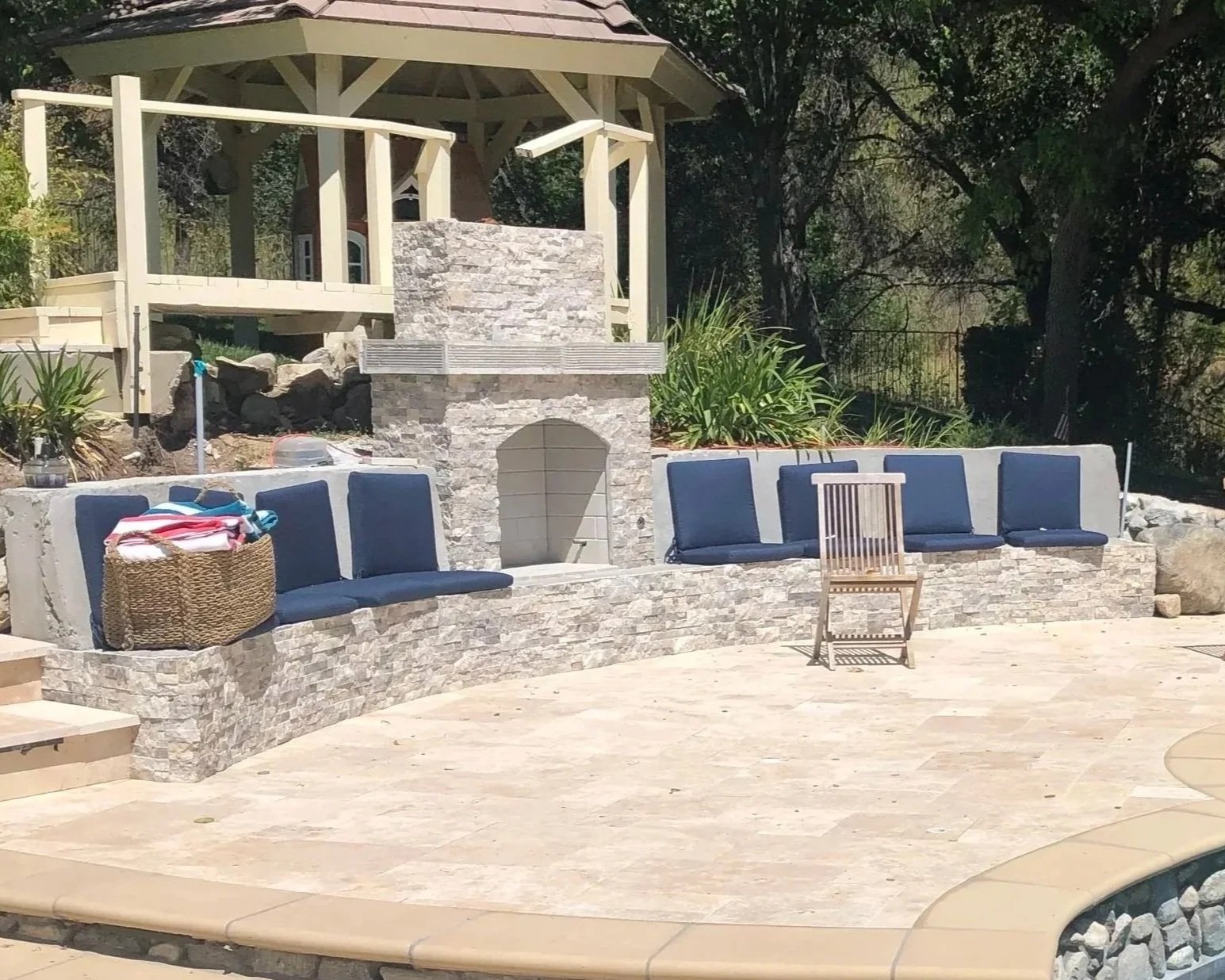 Backyard patio with a built-in stone firepit, blue cushioned chairs, a wooden chair, and a gazebo in the background surrounded by trees and plants.
