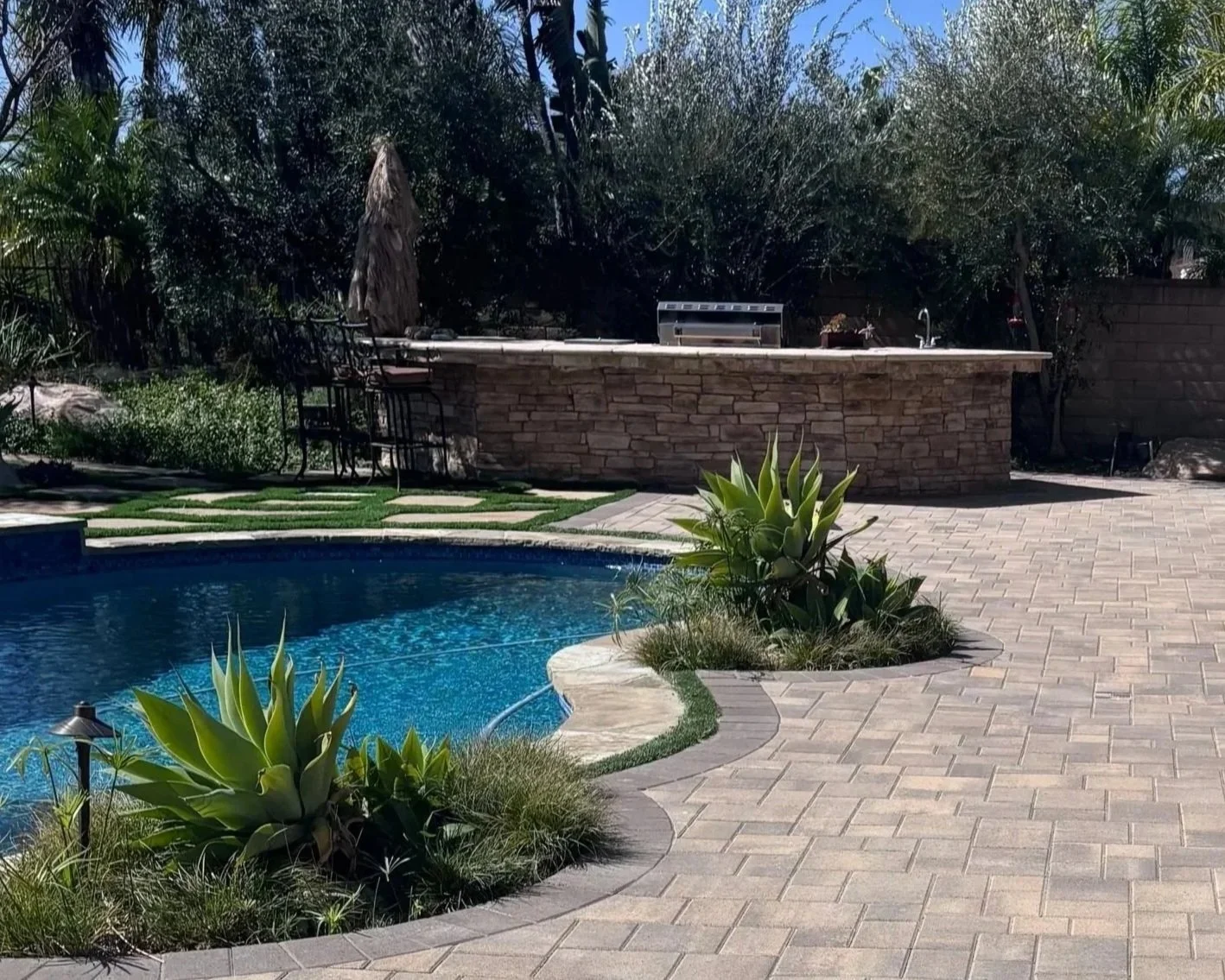 A backyard with a kidney-shaped swimming pool, surrounded by potted plants and paved with brick. There is a raised stone counter with a built-in grill and an outdoor faucet in the background, with greenery and trees beyond.