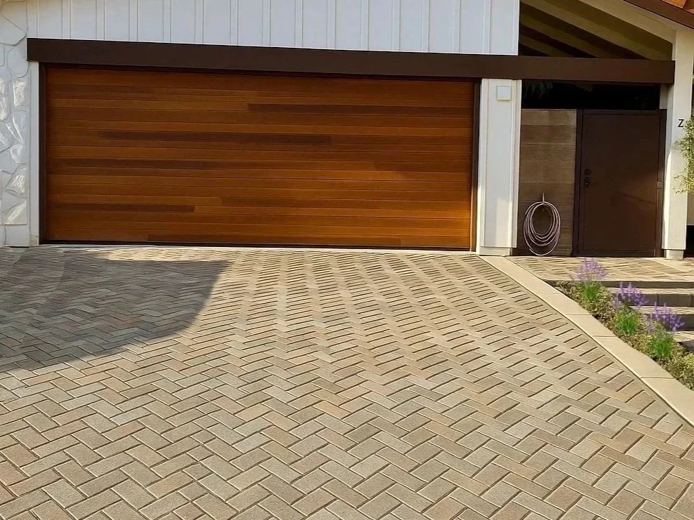A brick driveway leading to a garage with a wooden door, next to a small garden with purple flowers and a black door.