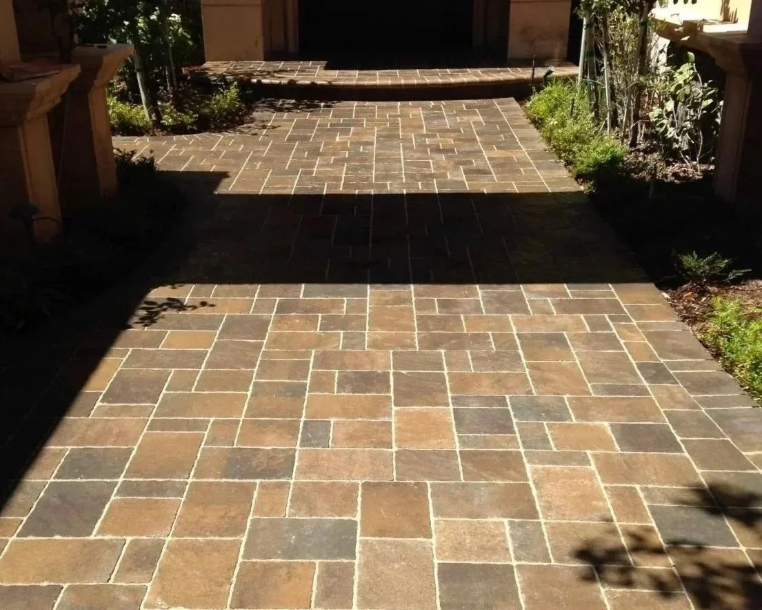 A brick or stone paved outdoor patio area with some greenery along the sides and steps leading up to a door.