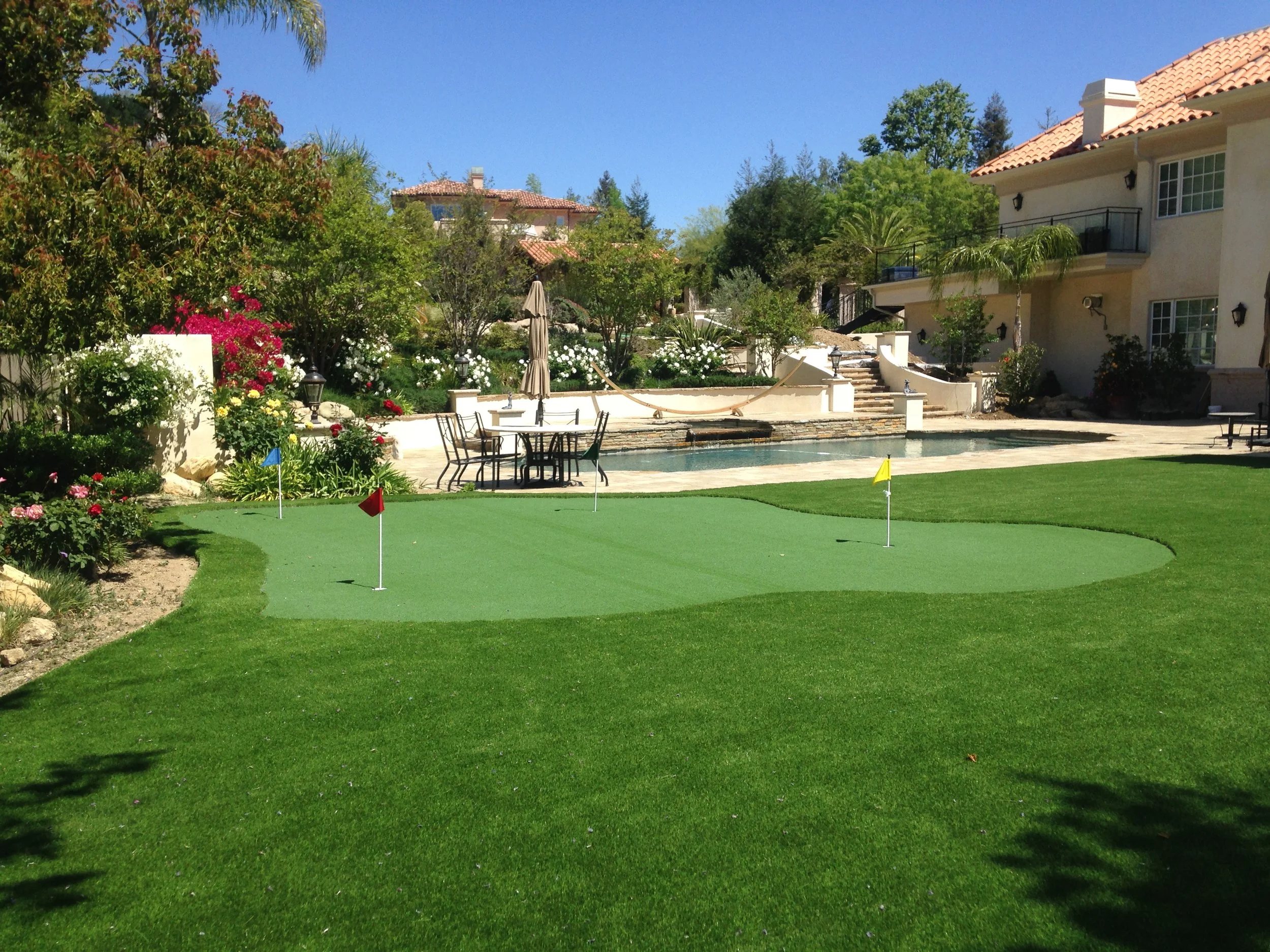 A backyard with a putting green, a pool with a winding water feature, surrounded by lush greenery and flowering plants, with a house in the background.
