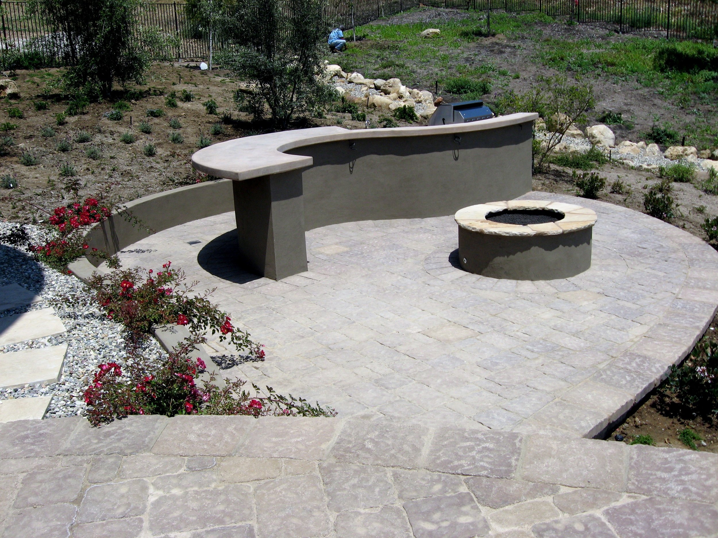 A backyard garden with a concrete seating area, a fire pit, a planter with pink flowers, and a hillside with various plants behind a black metal fence.