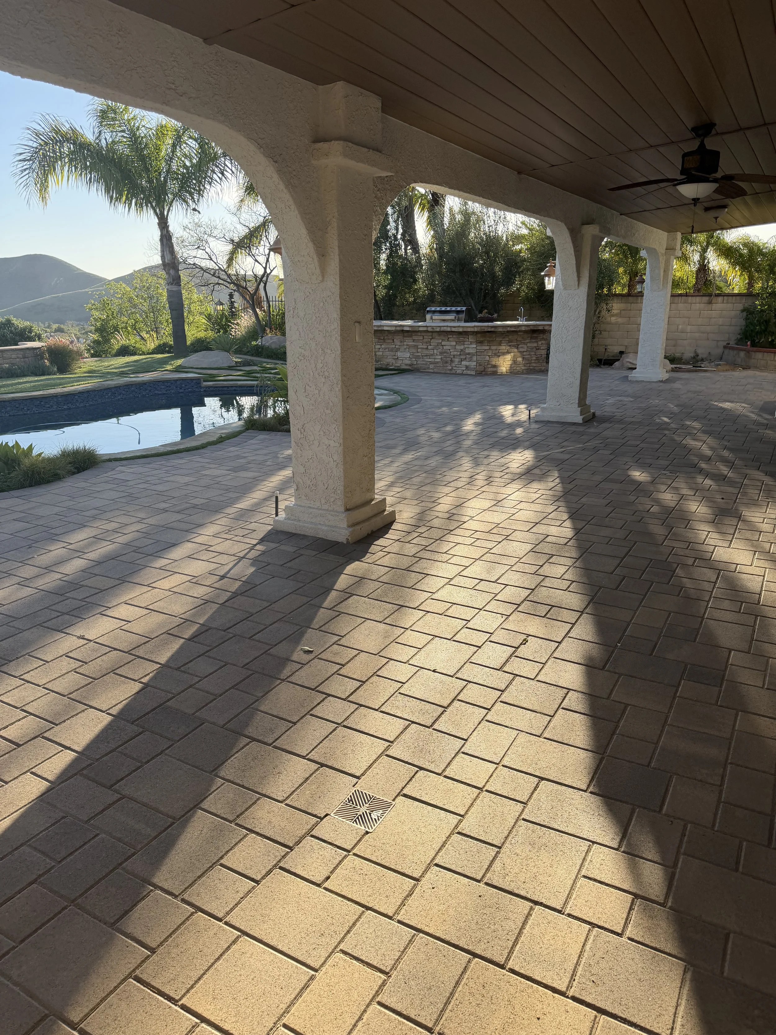 Patio area with beige brick pavers, overlooking a pool and lush green landscaping with trees and hills in the background, shaded by a wooden ceiling with ceiling fans.