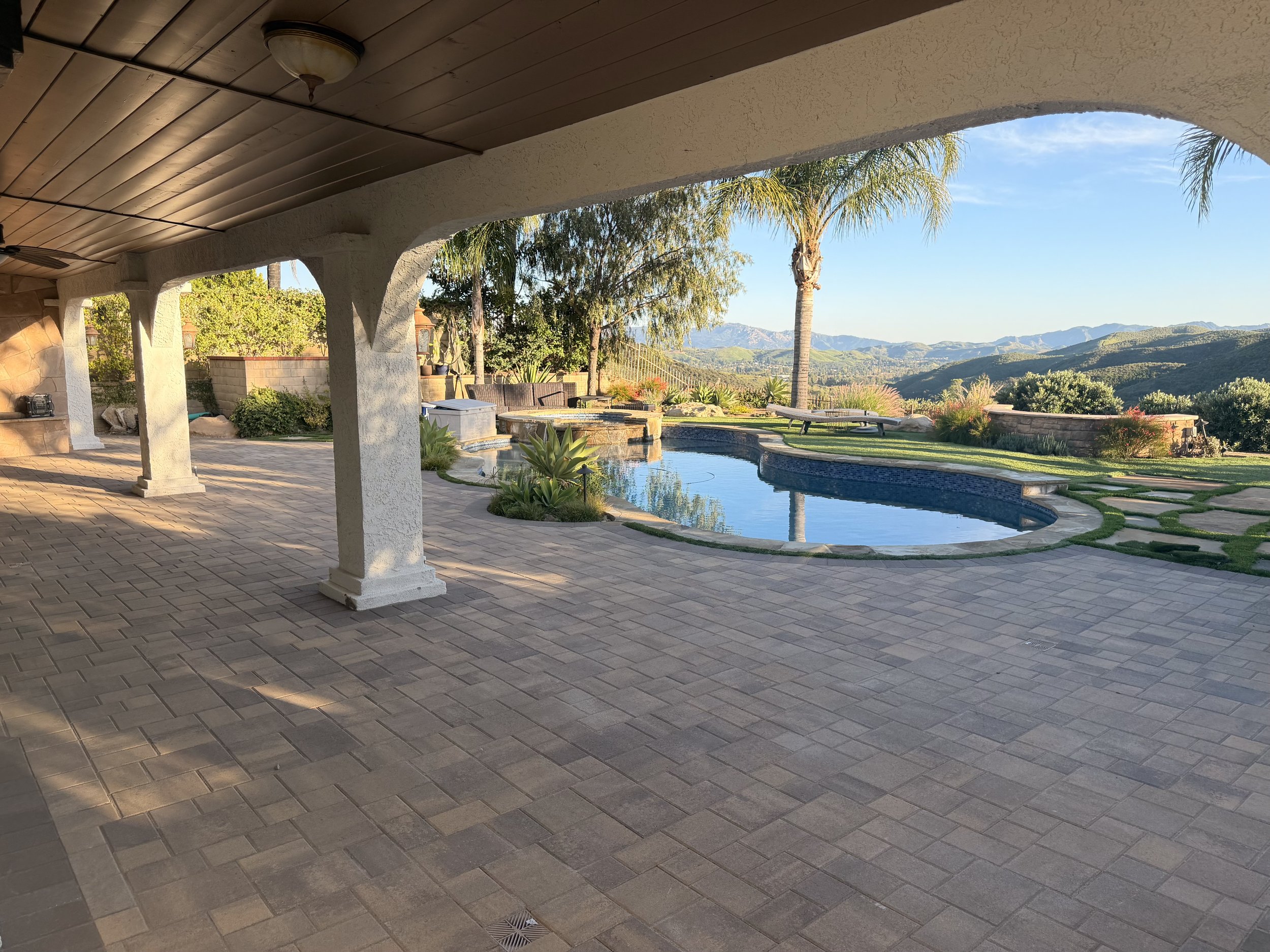 An outdoor patio area with a covered roof, brick flooring, a swimming pool, and scenic mountains in the background, along with palm trees and outdoor furniture.