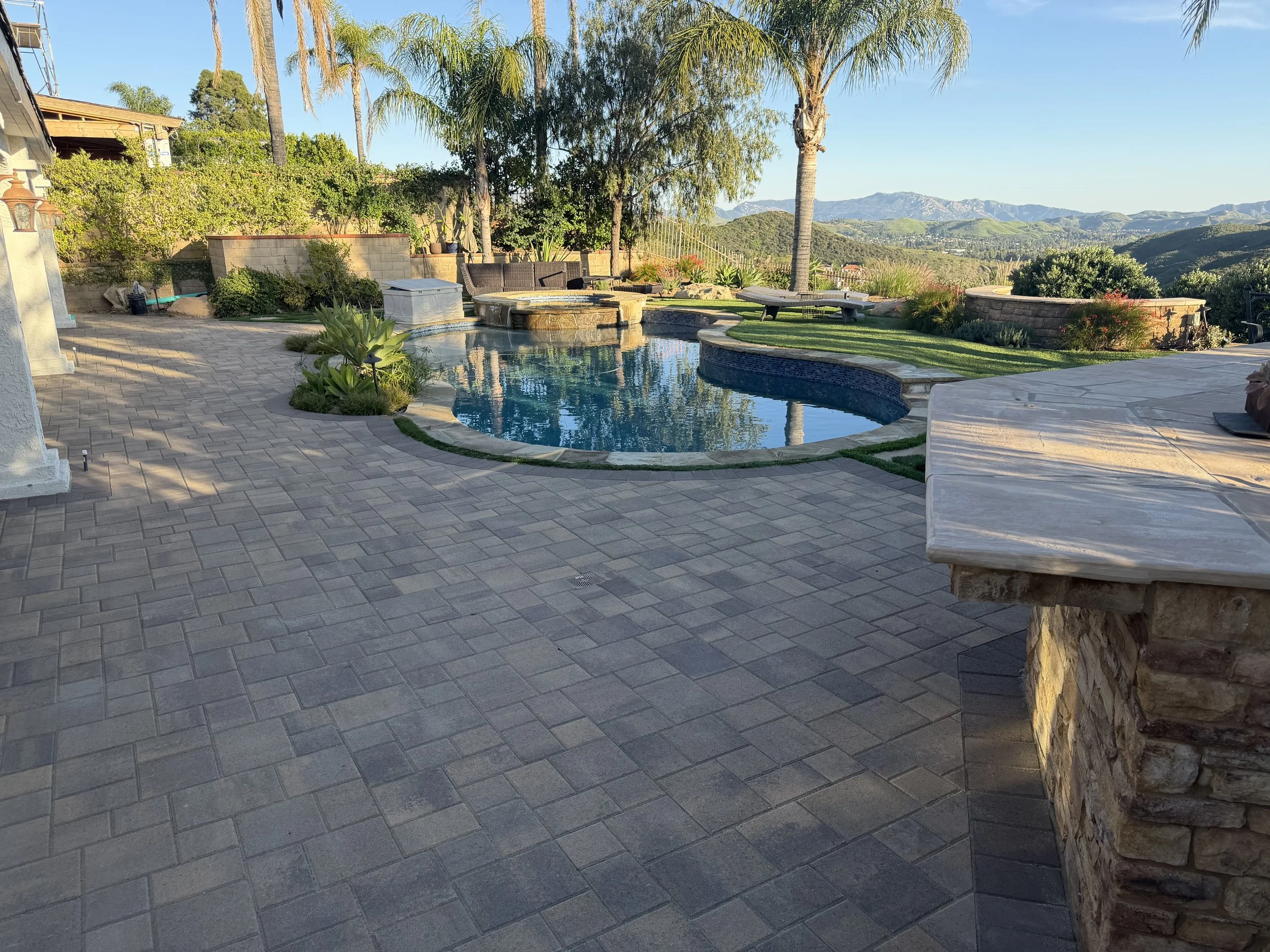 Outdoor backyard scene with a swimming pool, lounge chairs, trees, and a scenic mountain view in the background during daylight.