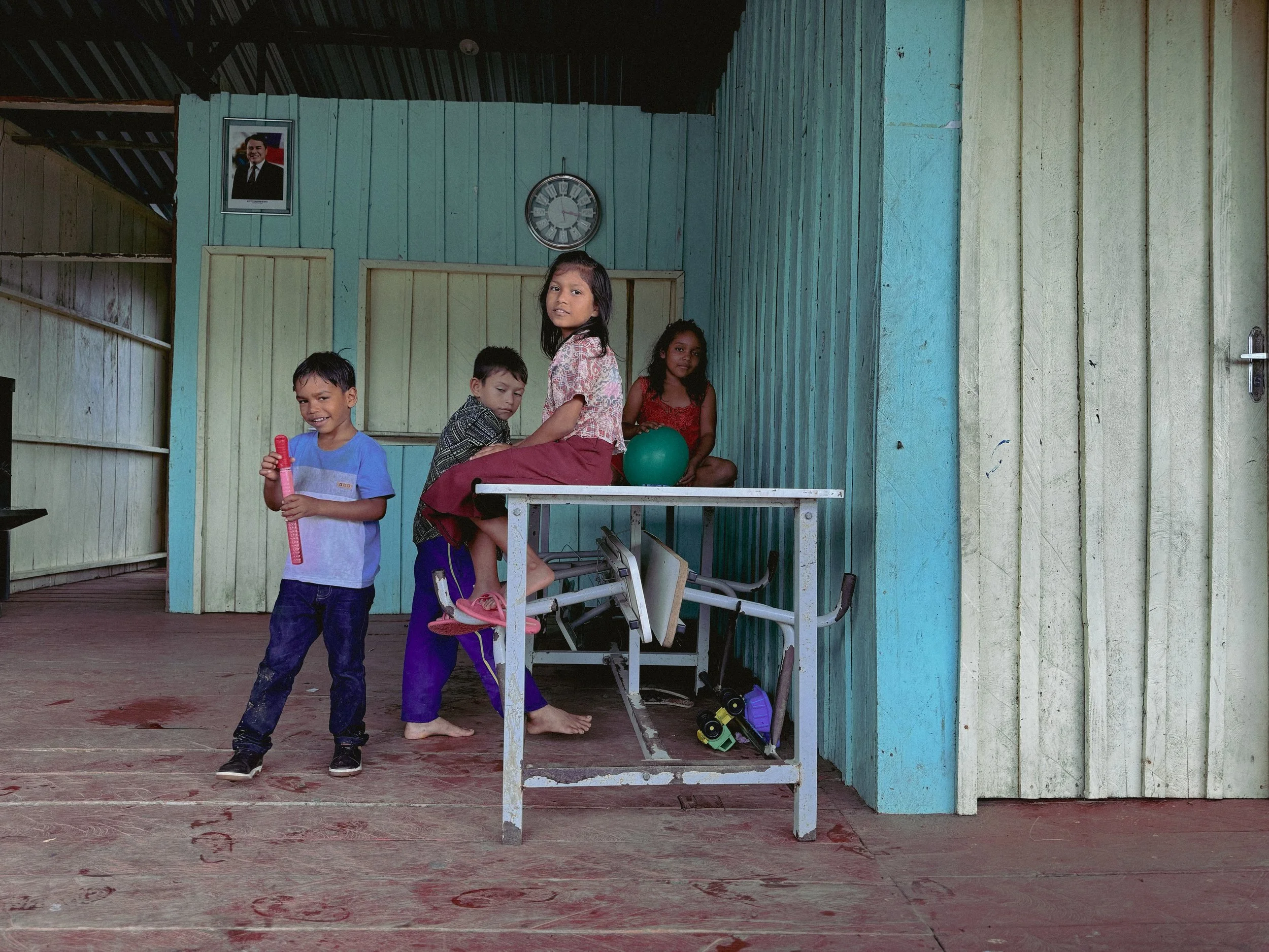 Four children and a woman in a blue wooden house with a clock on the wall, a framed photo, and a table with a green ball, with toys on the floor.