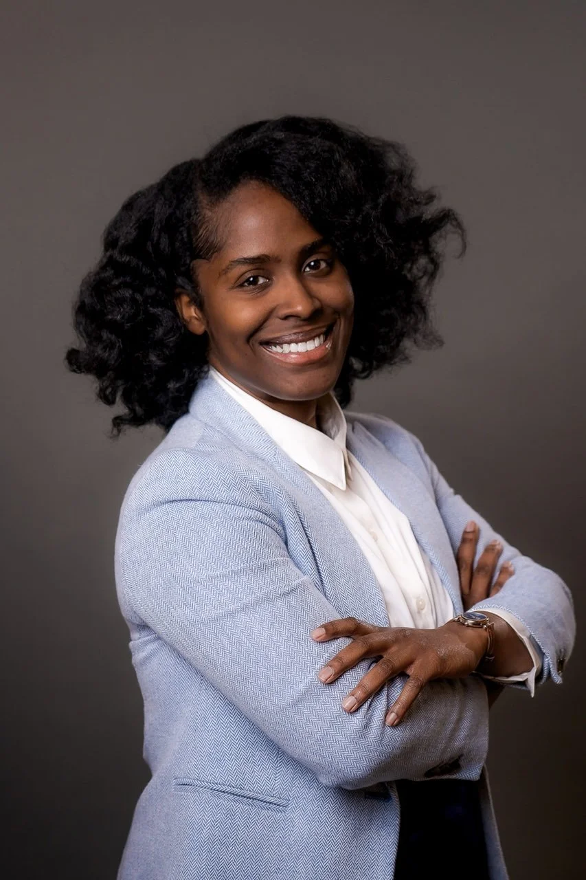 A professional woman with dark curly hair and a broad smile, wearing a light blue blazer and white shirt, standing with her arms crossed against a gray background.
