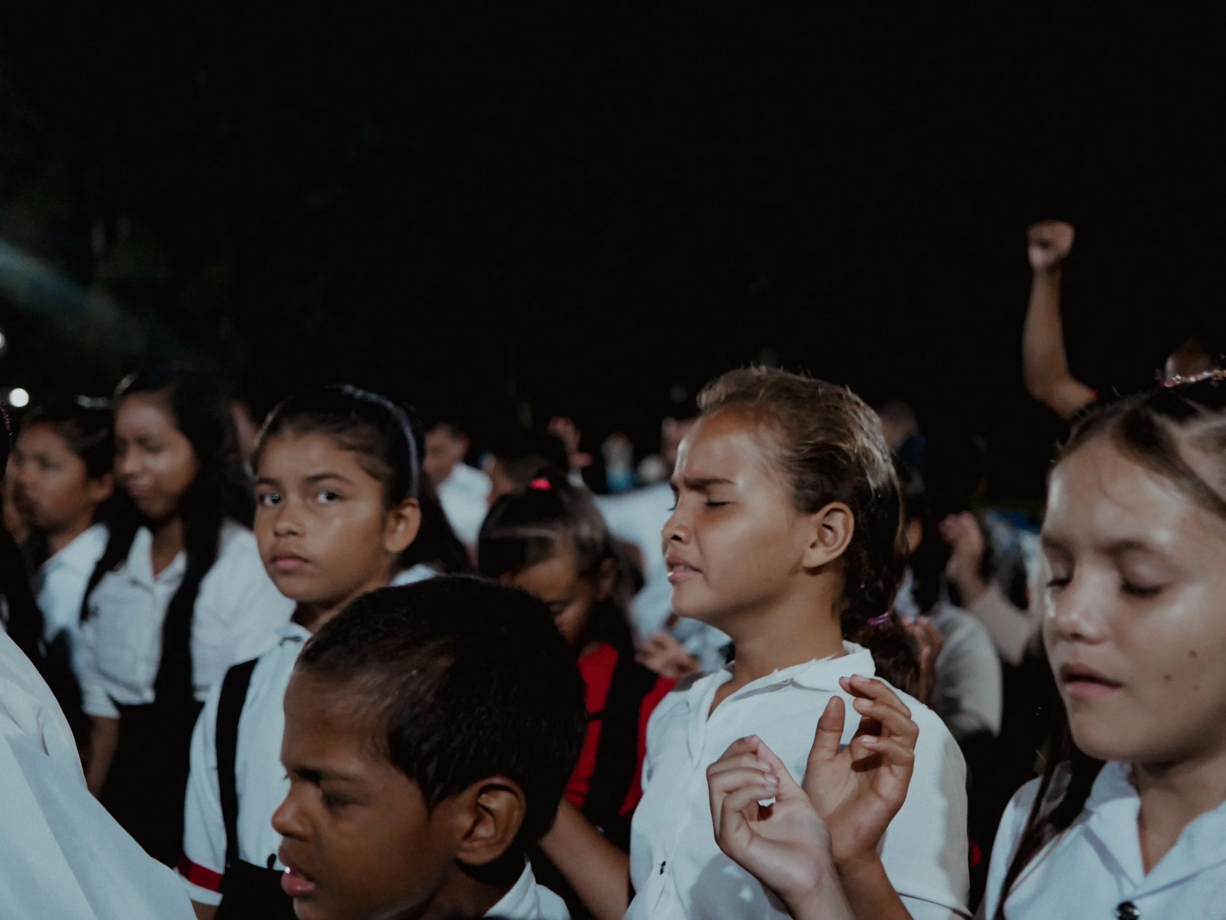A group of children dressed in martial arts uniforms, standing closely together at night, some with eyes closed and one with a raised hand.