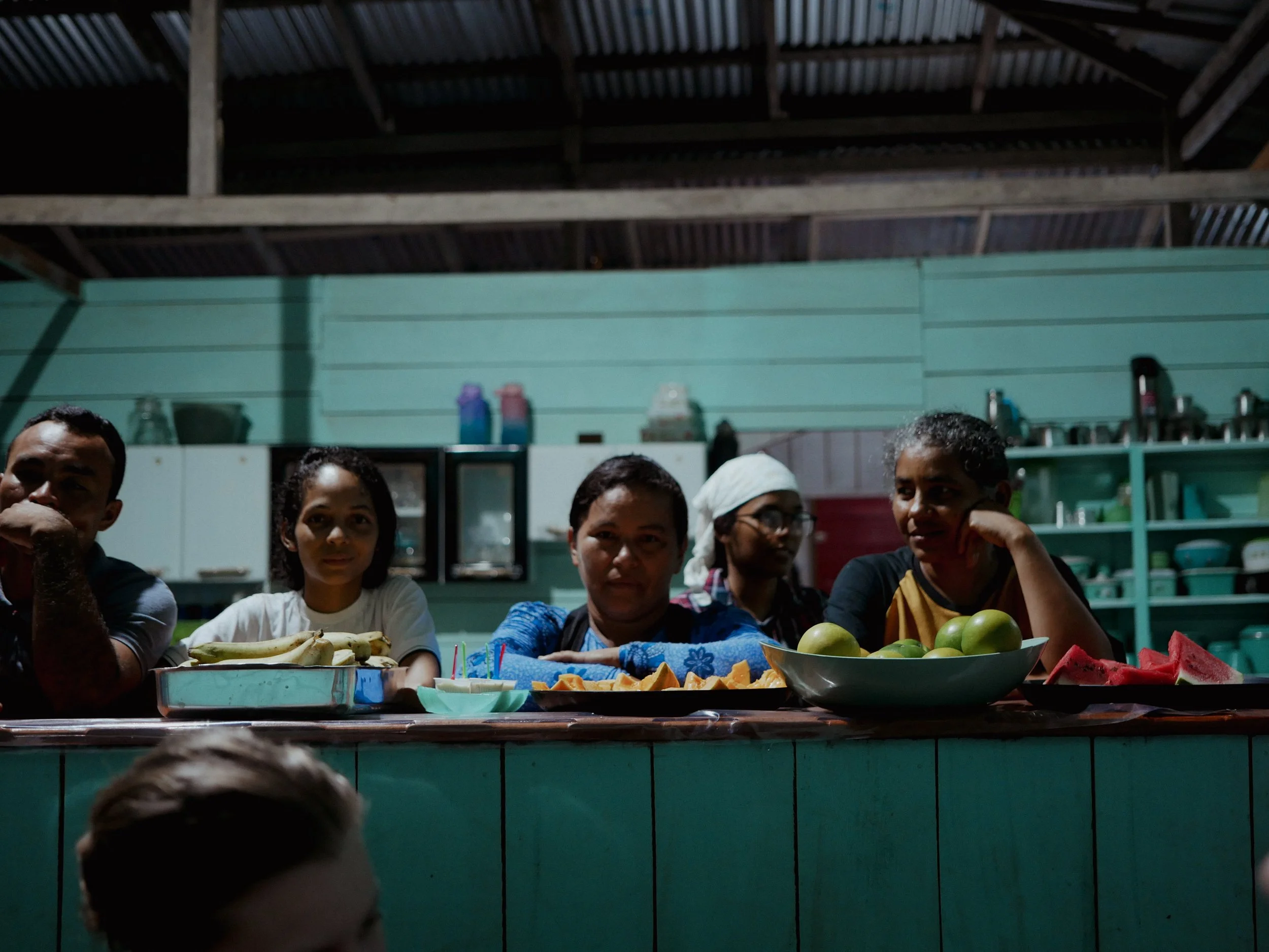 People sitting at a table with fruit dishes, including bananas, watermelon, apples, and sliced papaya, inside a room with a green wall and metal roof.