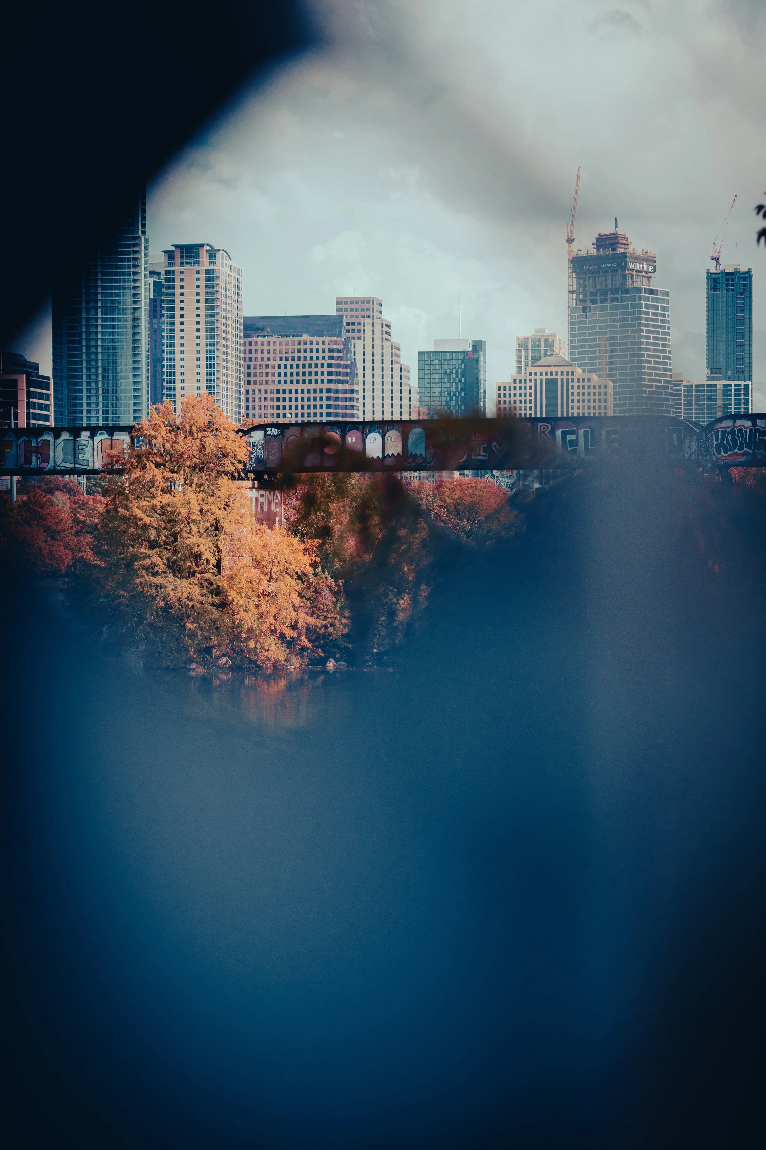 A city skyline with modern skyscrapers, seen through a through a small opening or frame, with some graffiti-covered train tracks in the foreground and autumn-colored trees near a body of water.