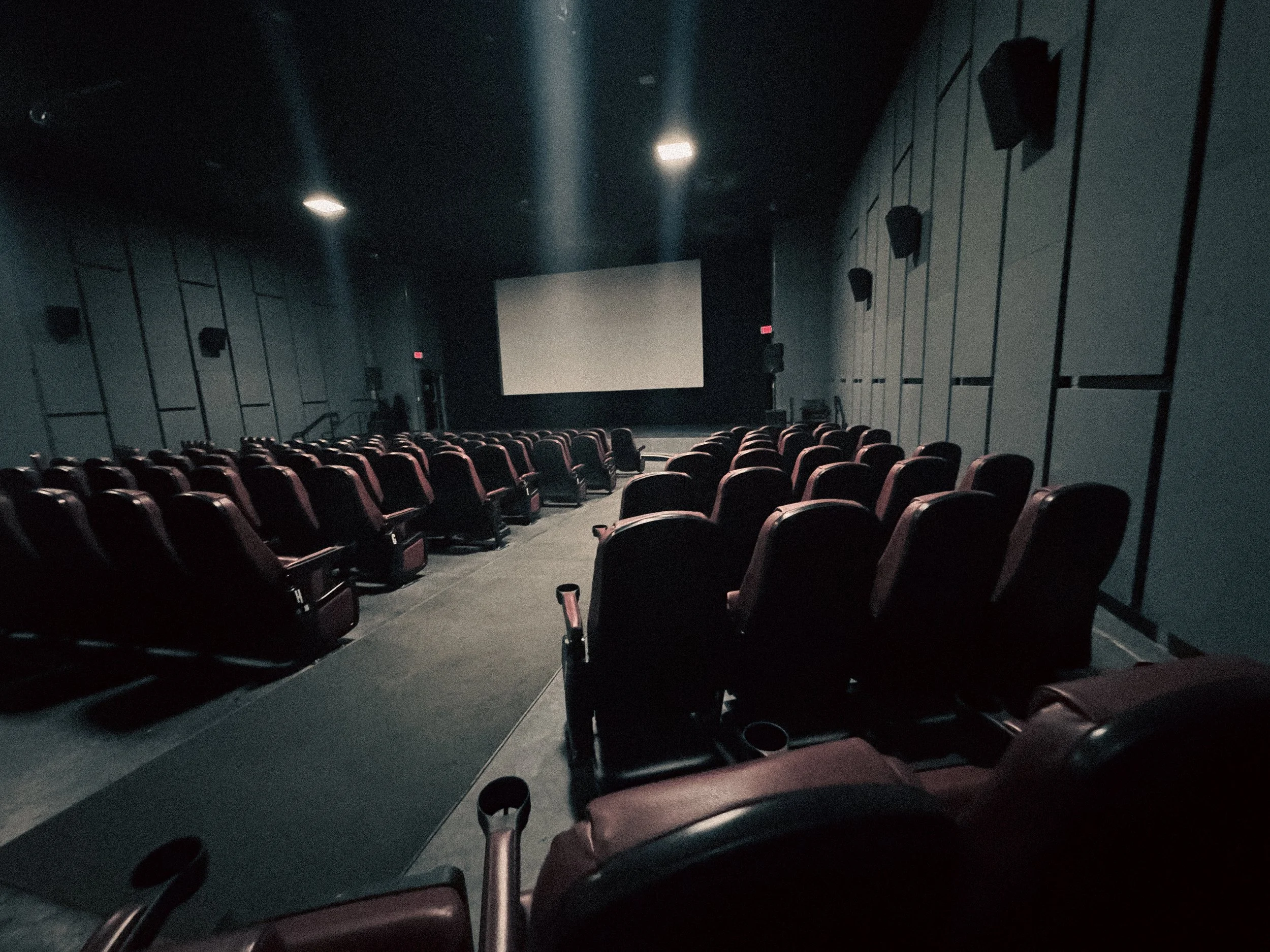 An empty movie theater with rows of red theater seats facing a large blank screen, dim lighting, and dark walls.