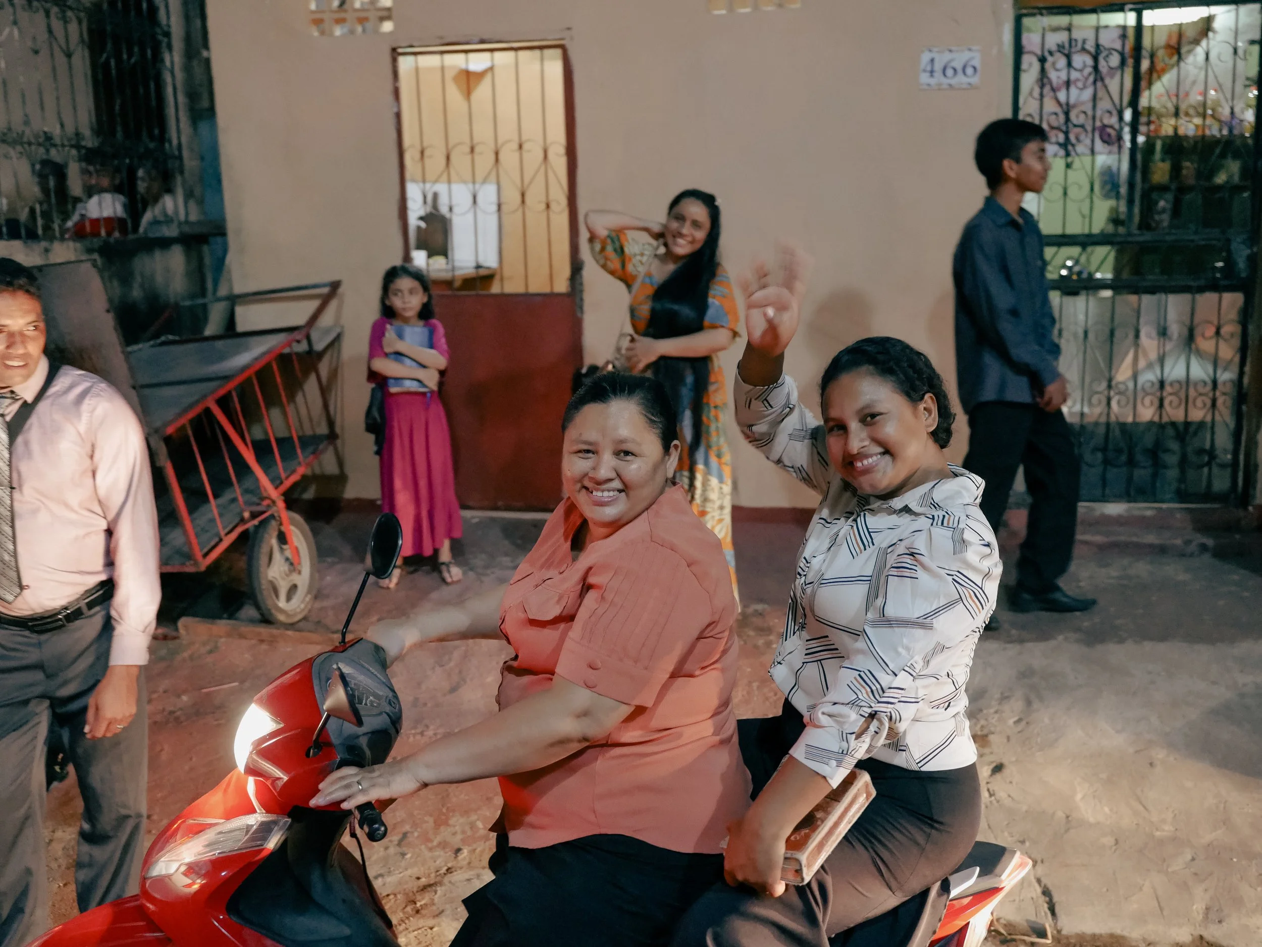 A group of people gathered outside a building at night, with some smiling at the camera; a woman in a peach shirt is sitting on a motor scooter, and a woman in a striped shirt is standing beside her.