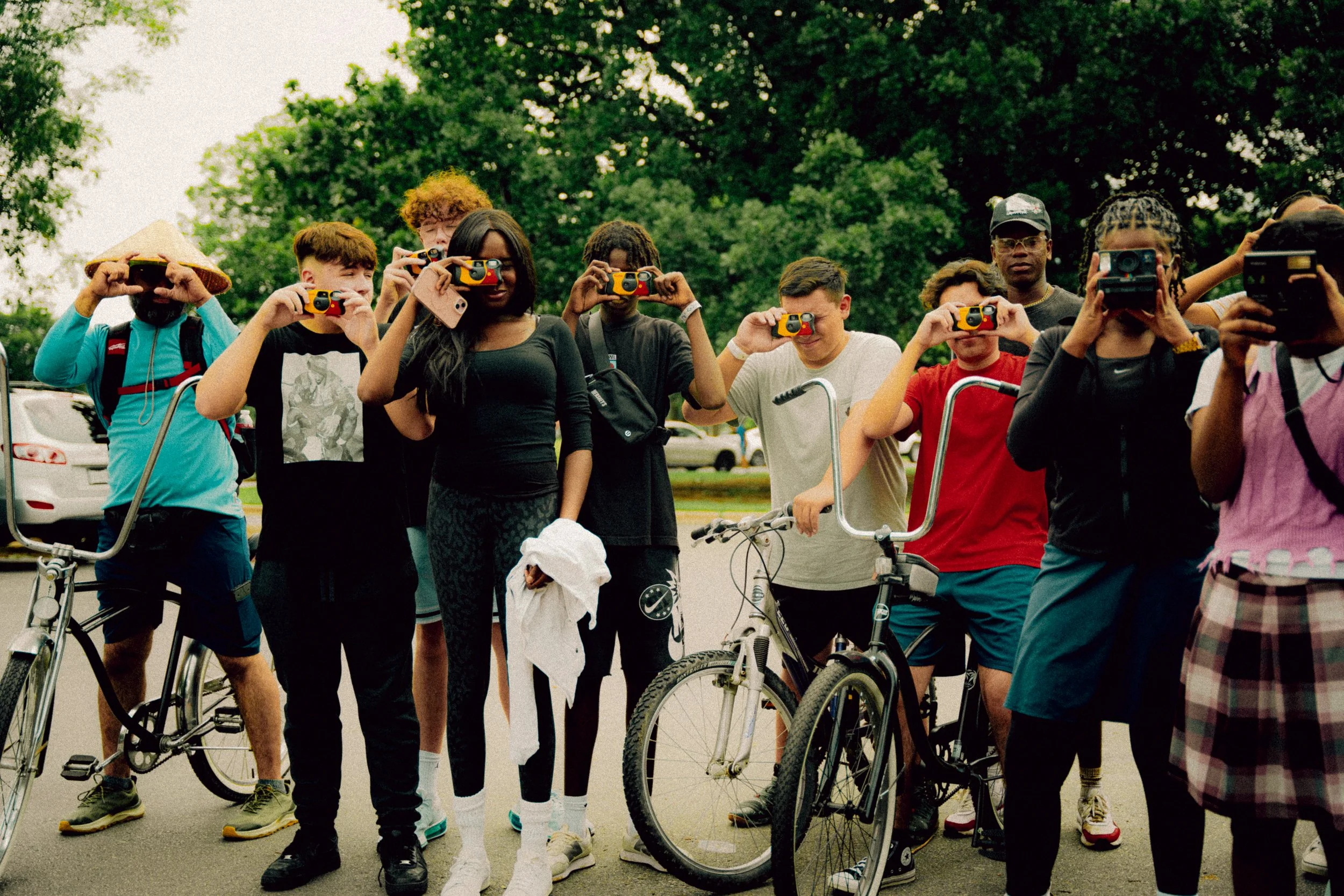 A group of diverse young people standing outdoors, taking photos with their cameras and holding bicycles.