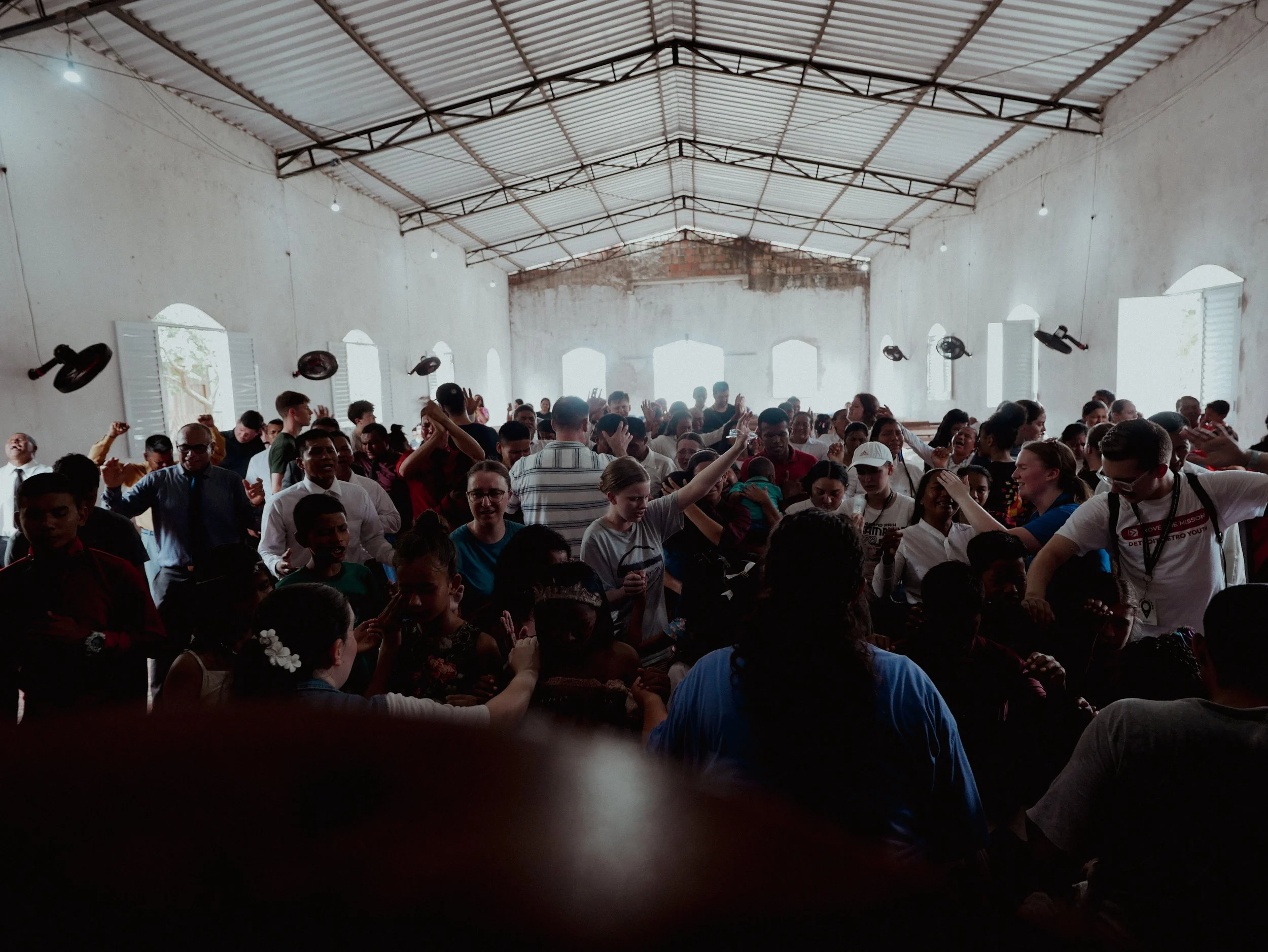 A large group of people gathered inside a spacious building with white walls and a metal roof. The crowd appears to be enjoying an event, with many raising their hands or dancing. Light comes in through open windows on both sides.