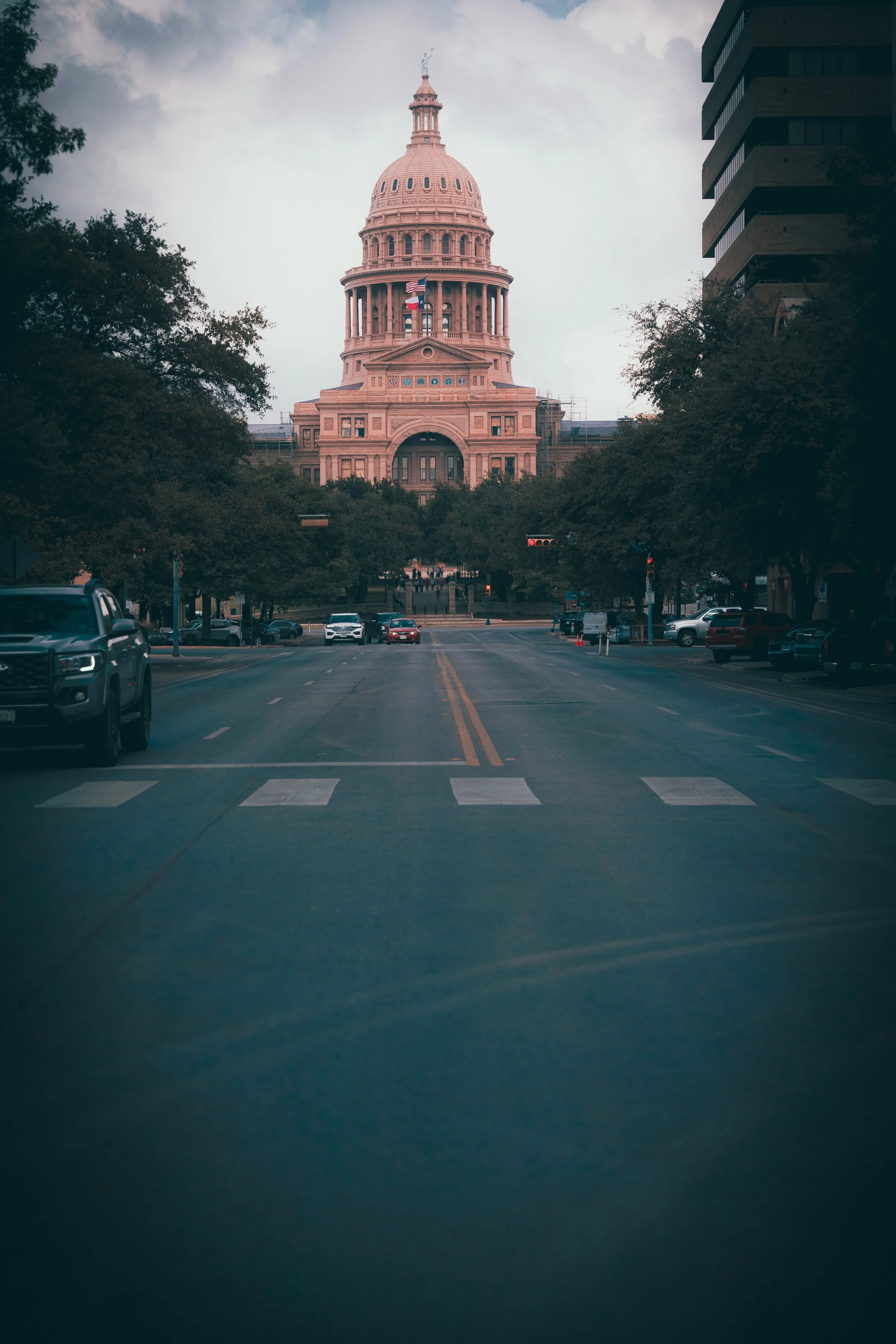 View of the Texas State Capitol in Austin during daytime, framed by trees on each side, cars on the street, and cloudy sky.