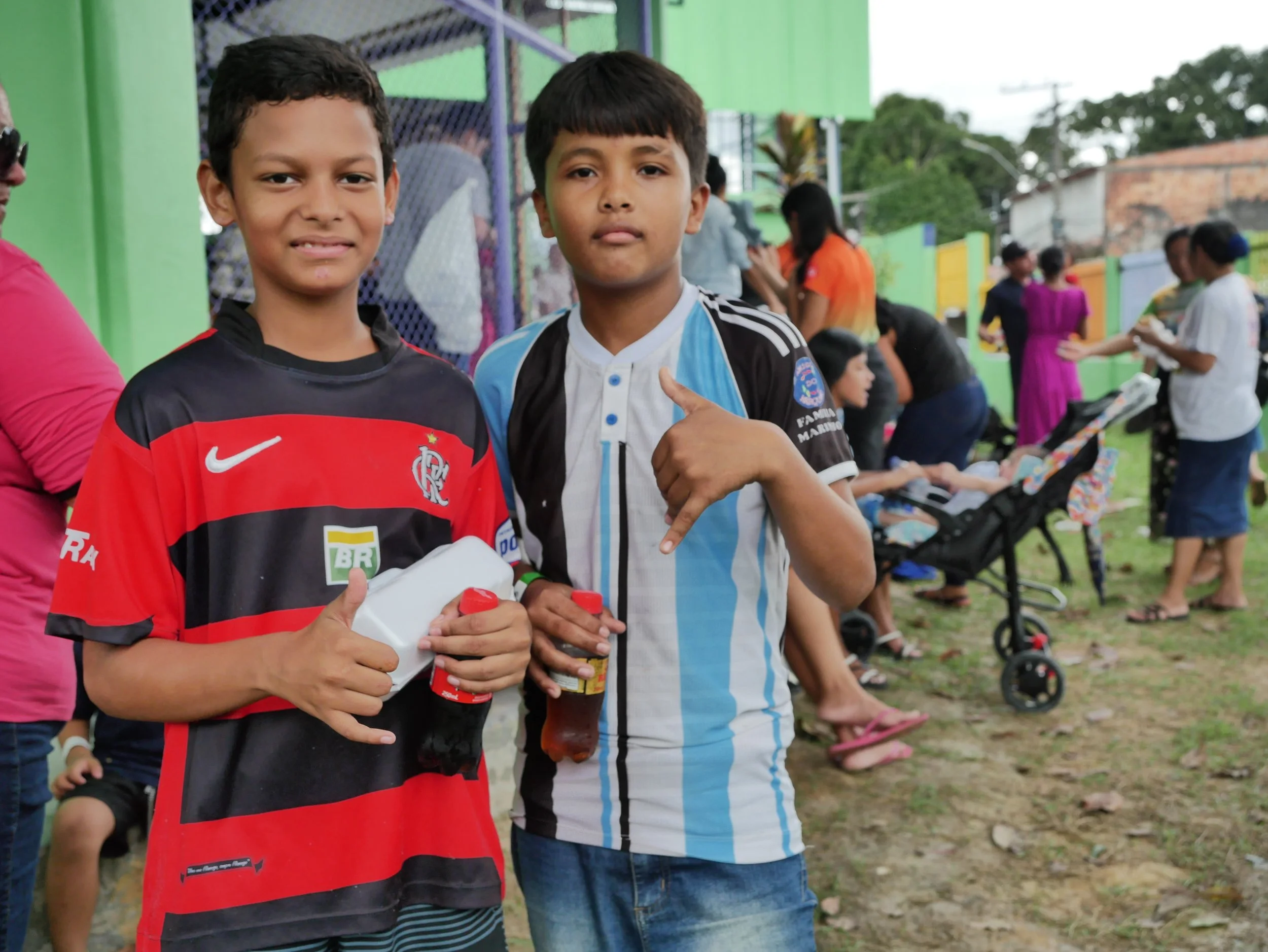 Two young boys standing outdoors, one wearing a red and black sports jersey and the other in a light blue and white striped soccer jersey, surrounded by people and strollers in the background.
