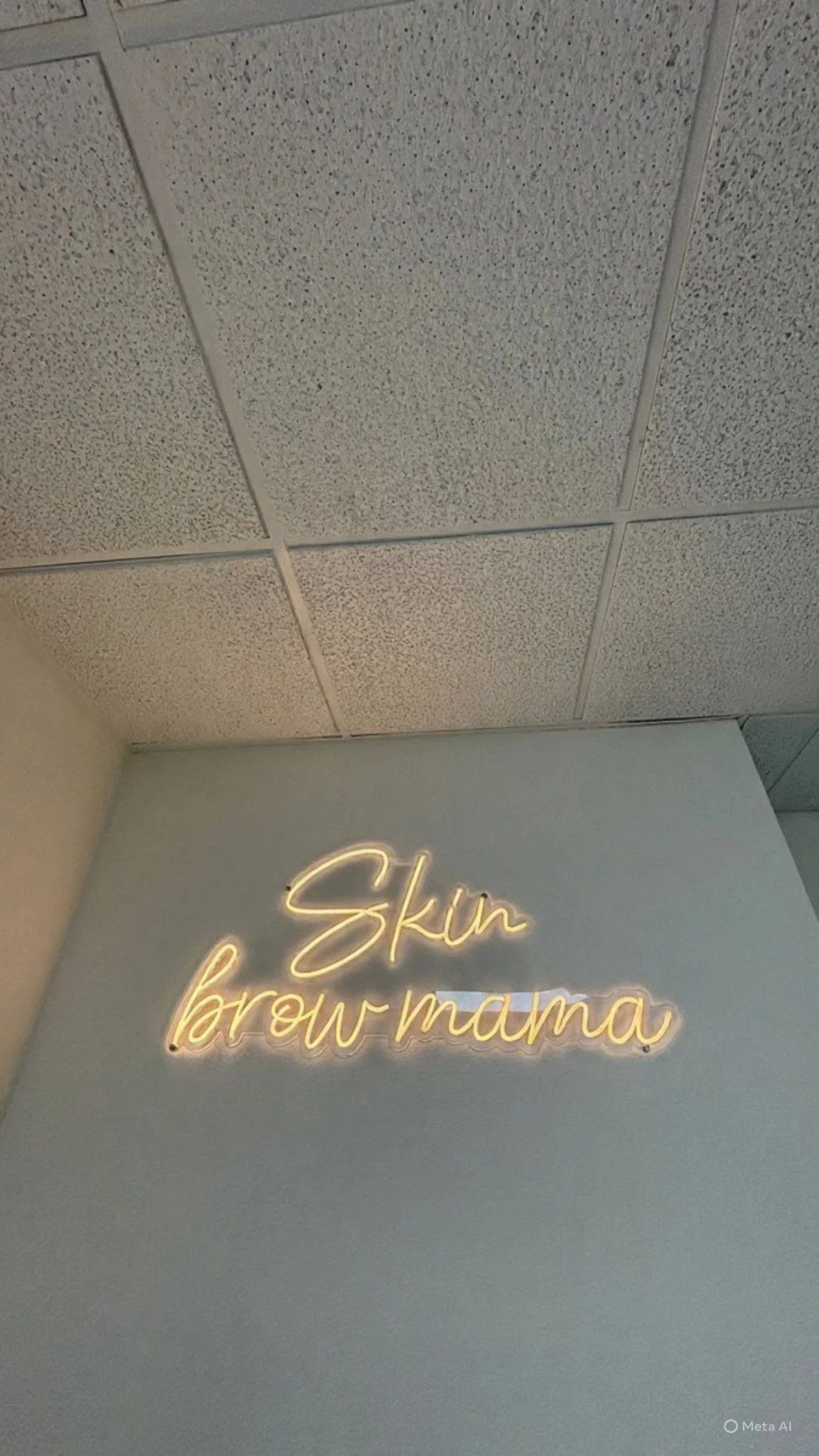 Neon sign reading 'Skin brow mama' on a light-colored wall beneath a textured ceiling.