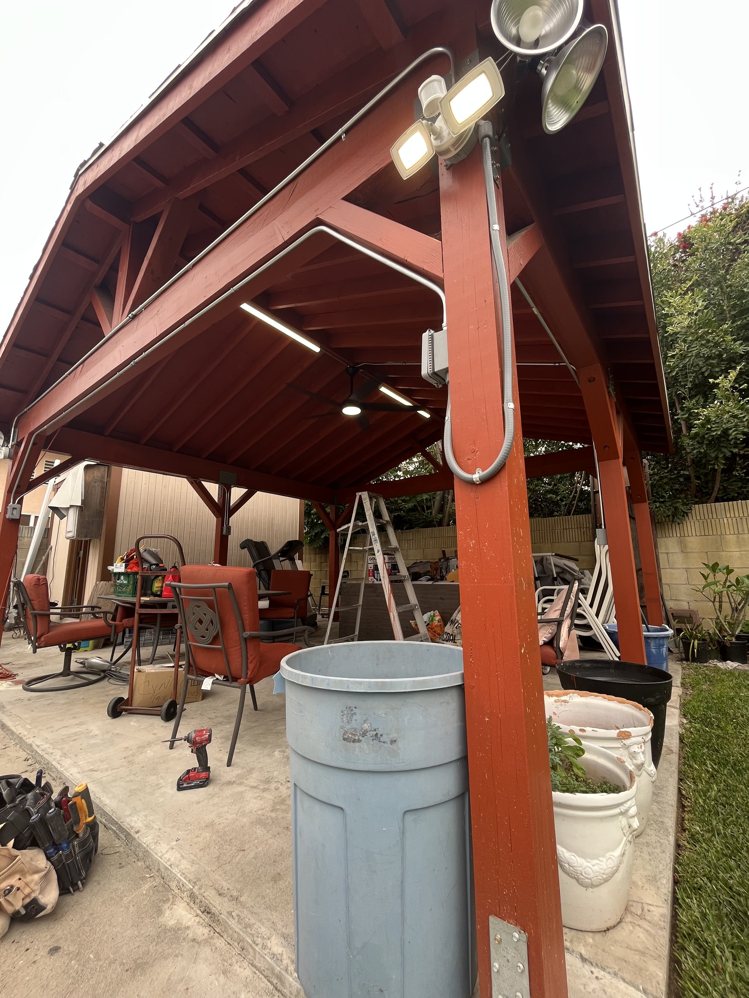 A backyard patio with a red wooden pergola, various outdoor chairs, a step ladder, tools, and gardening pots, some filled with plants, on a concrete surface.
