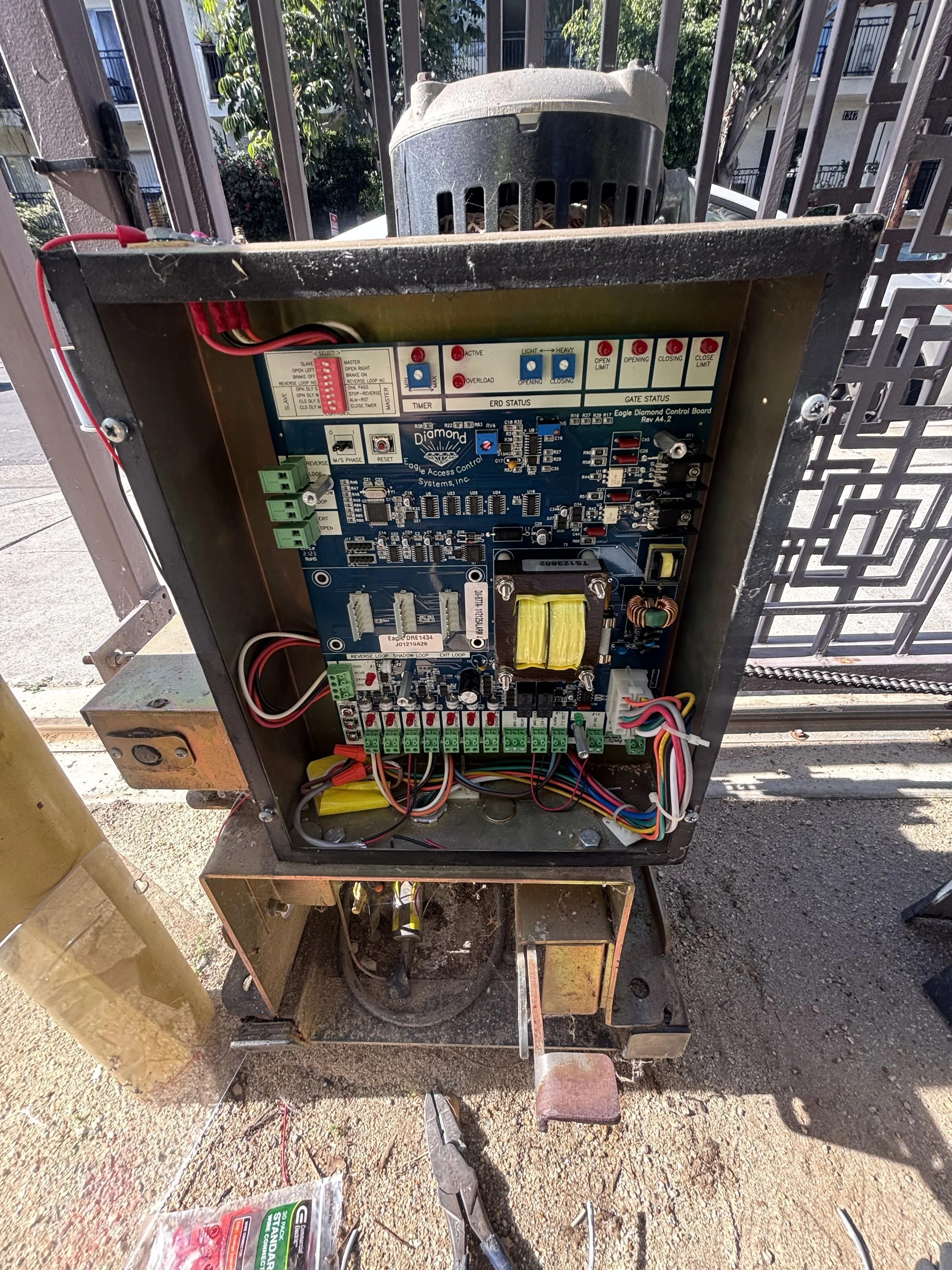 Open electronic gate control box with internal circuit board and wiring, mounted on a rusty metal frame outside on a sidewalk.
