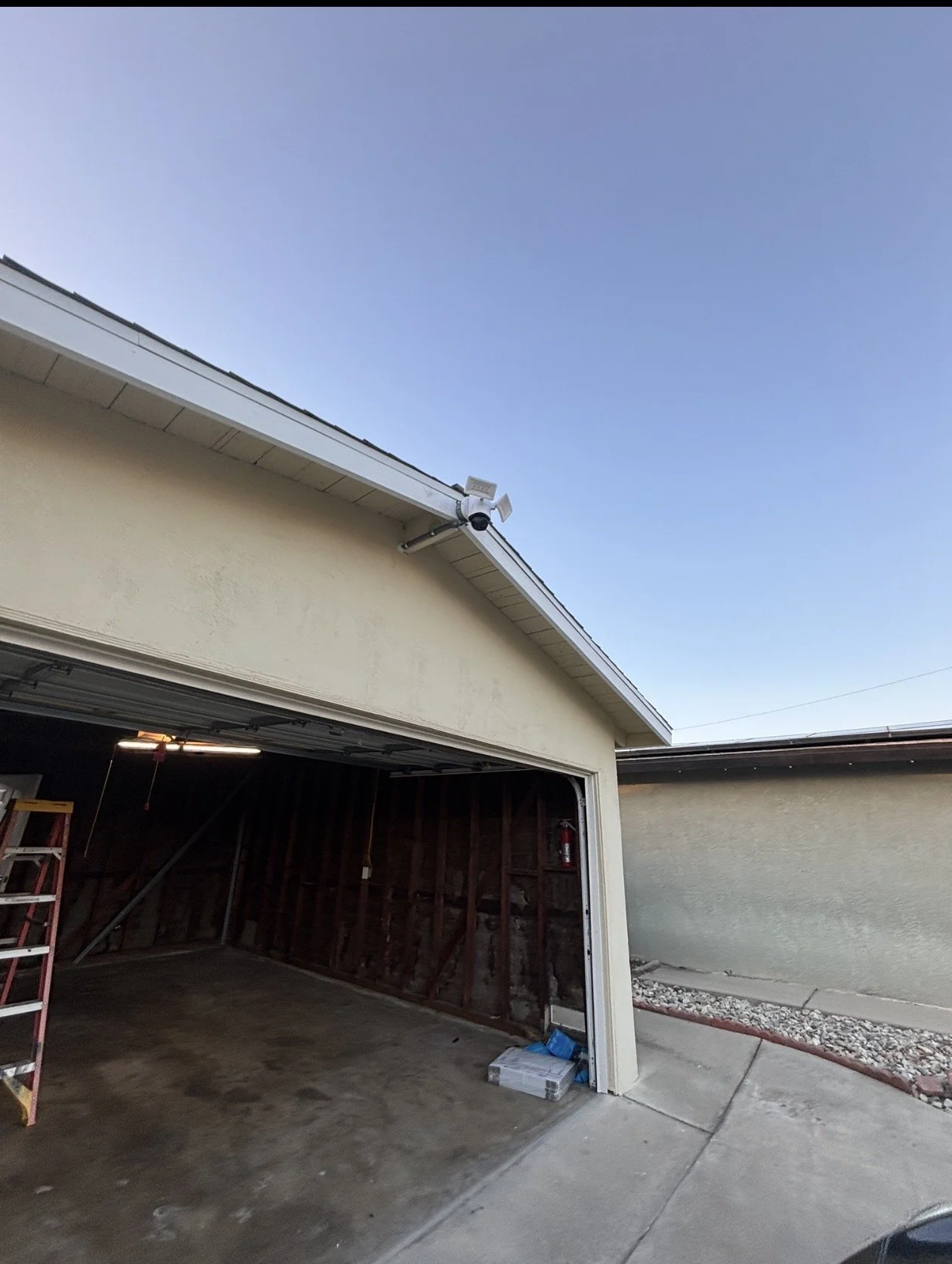 An open garage with a ladder inside, unfinished wall framing, a fire extinguisher on the side, and a clear sky overhead.
