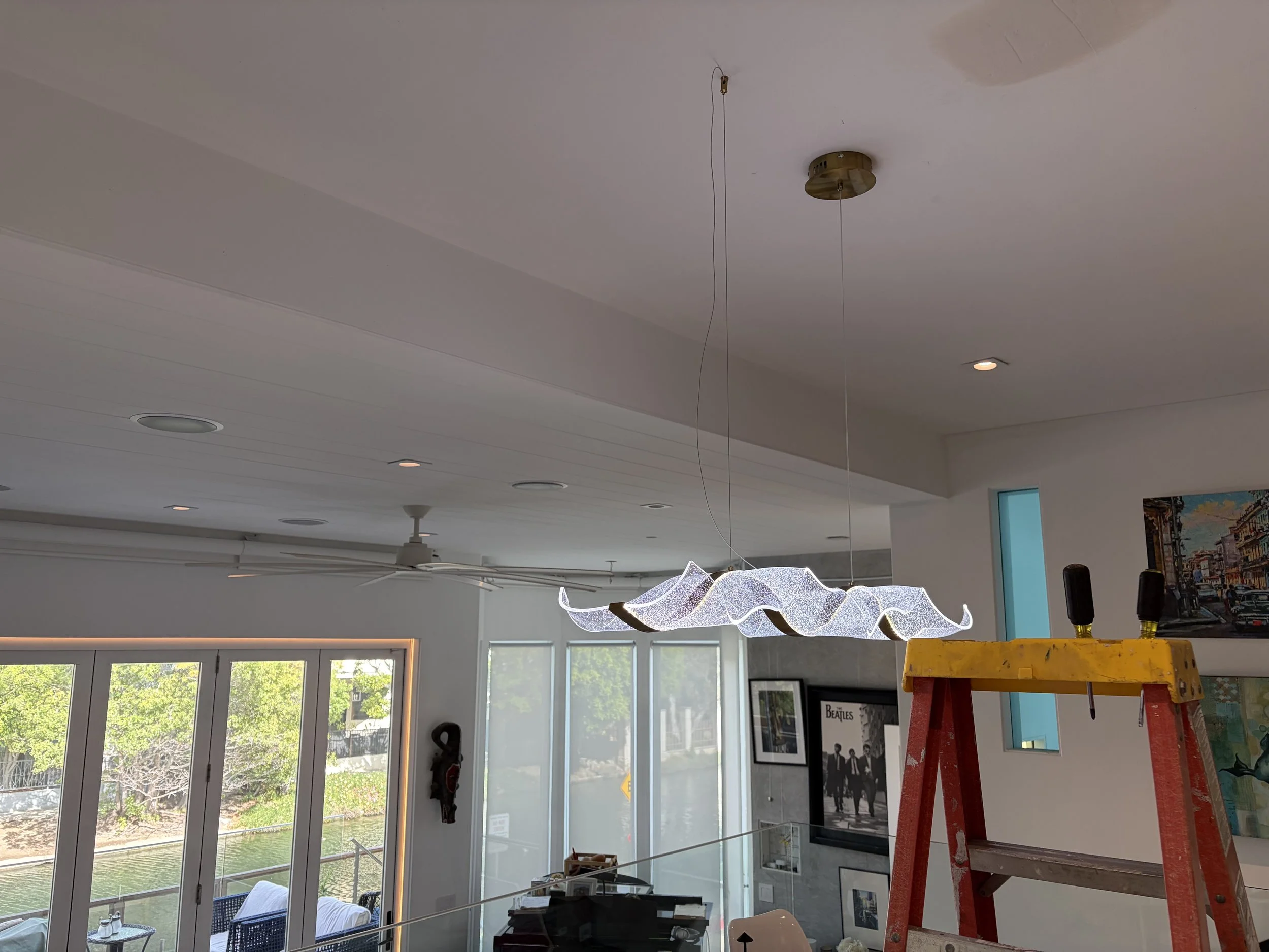 An art installation hanging from the ceiling with wavy illuminated elements. There is a yellow step ladder and a black painting of the Beatles on the wall.