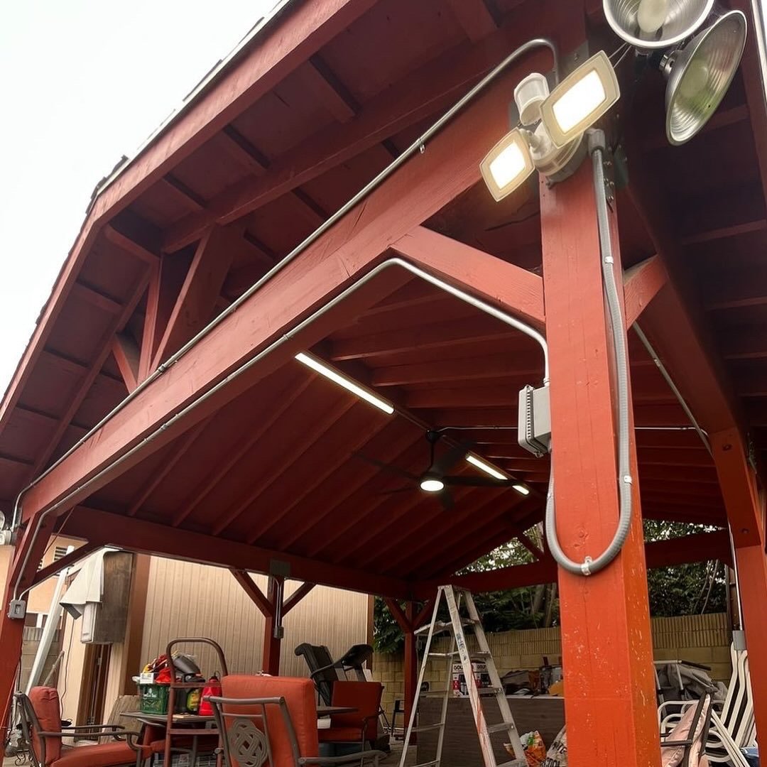 View of a covered outdoor area with wooden framework, ceiling lights, and various patio furniture and tools underneath, including a ladder and chairs.