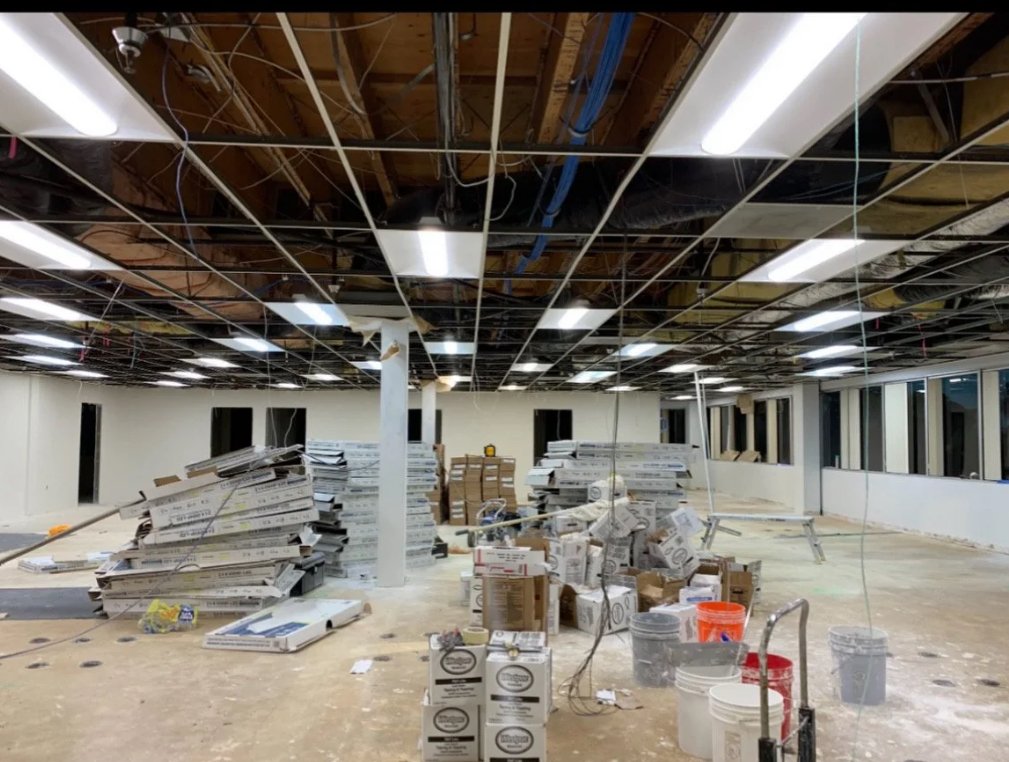 Room under construction with stacks of drywall and construction materials on the floor, exposed ceiling with plumbing and wiring, and windows along the wall.