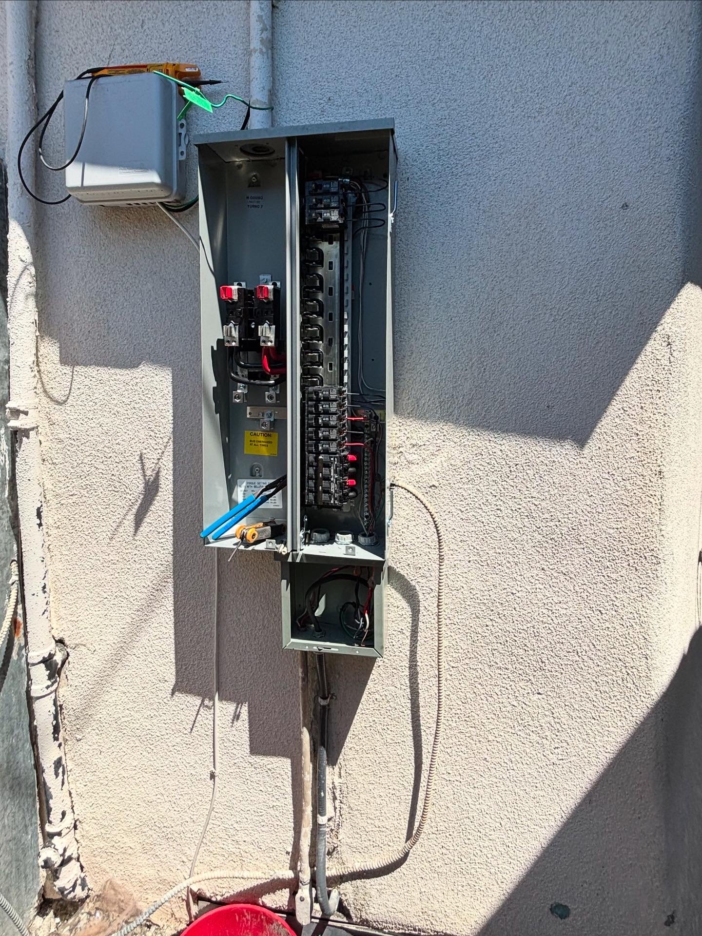 Outdoor electrical panel with open door showing circuit breakers and wiring mounted on a textured beige wall.
