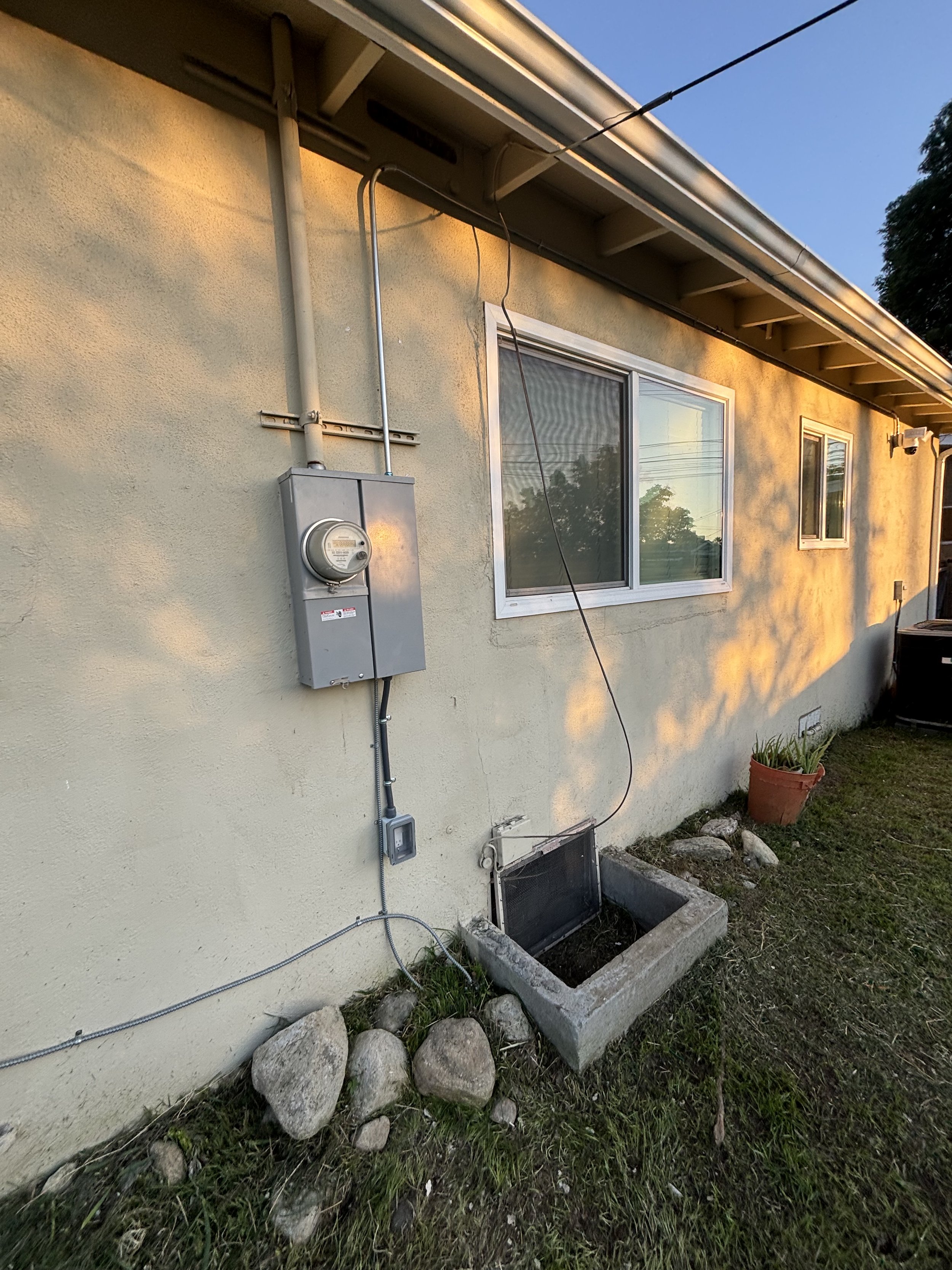 Side of a house with an electric meter, a window, a drain cover, a potted plant, and rocks at sunset.