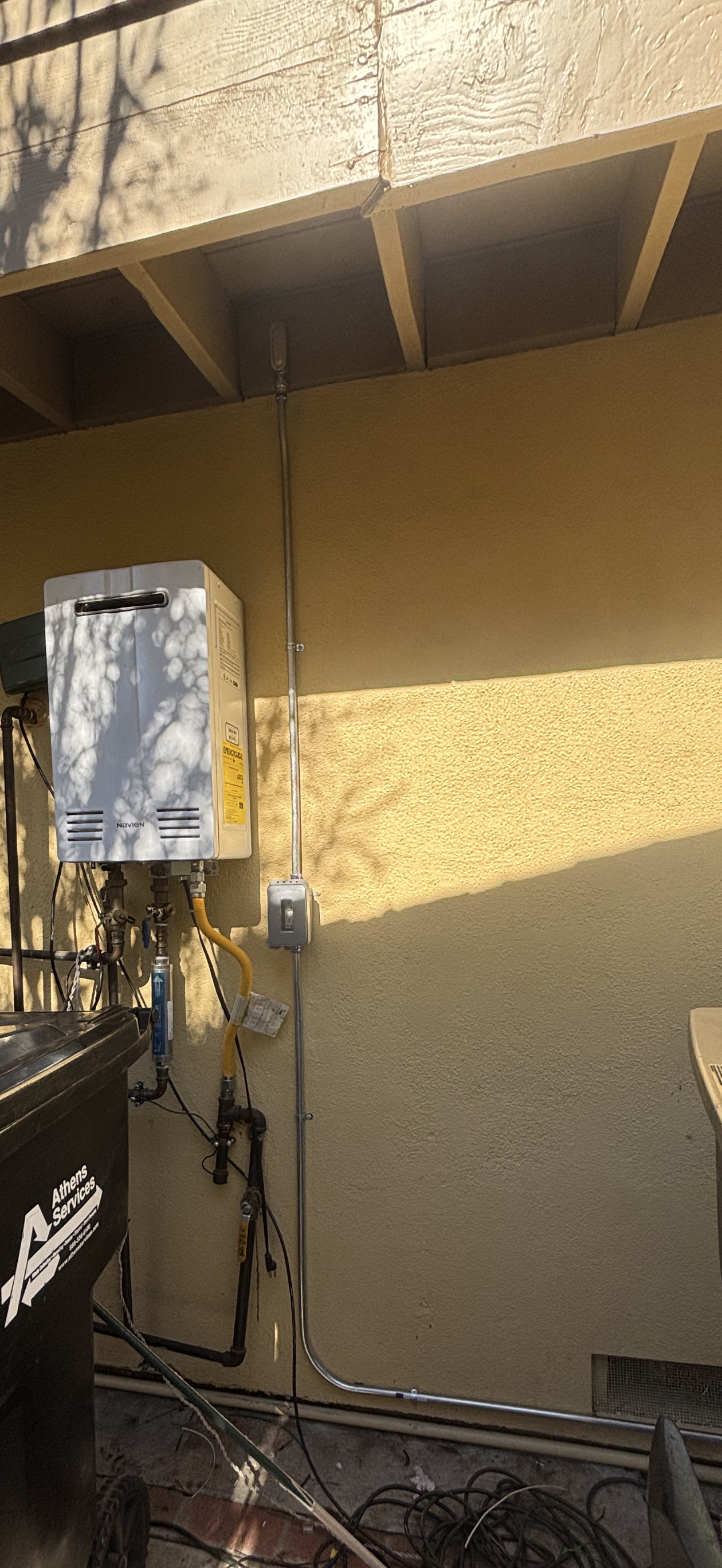 Exterior wall with an electrical water heater and plumbing pipes, with shadows of tree branches and sunlight on the wall.