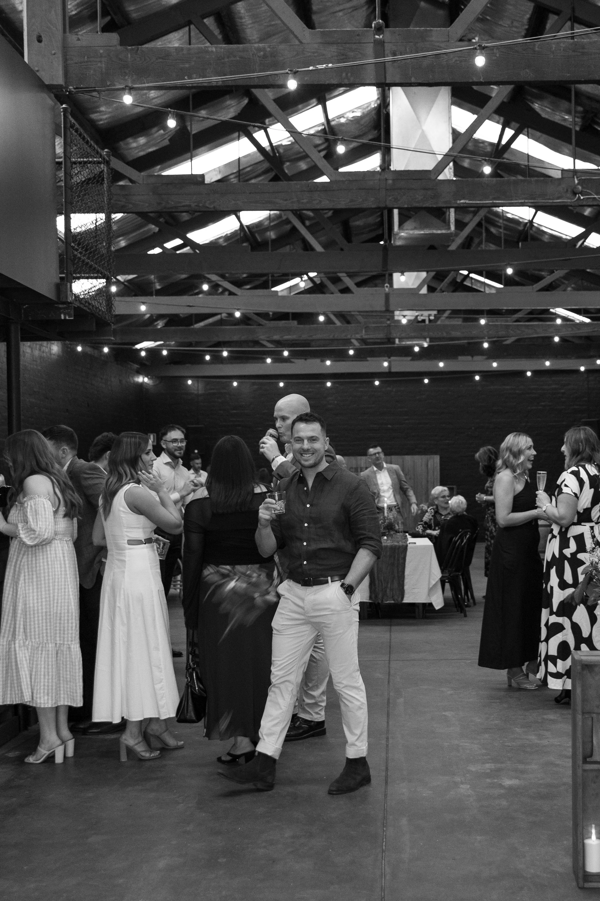 A group of people socializing at a formal event in a rustic, industrial-style venue with string lights hanging from the exposed wooden beams ceiling. The focus is on a man smiling and holding a drink in the foreground.