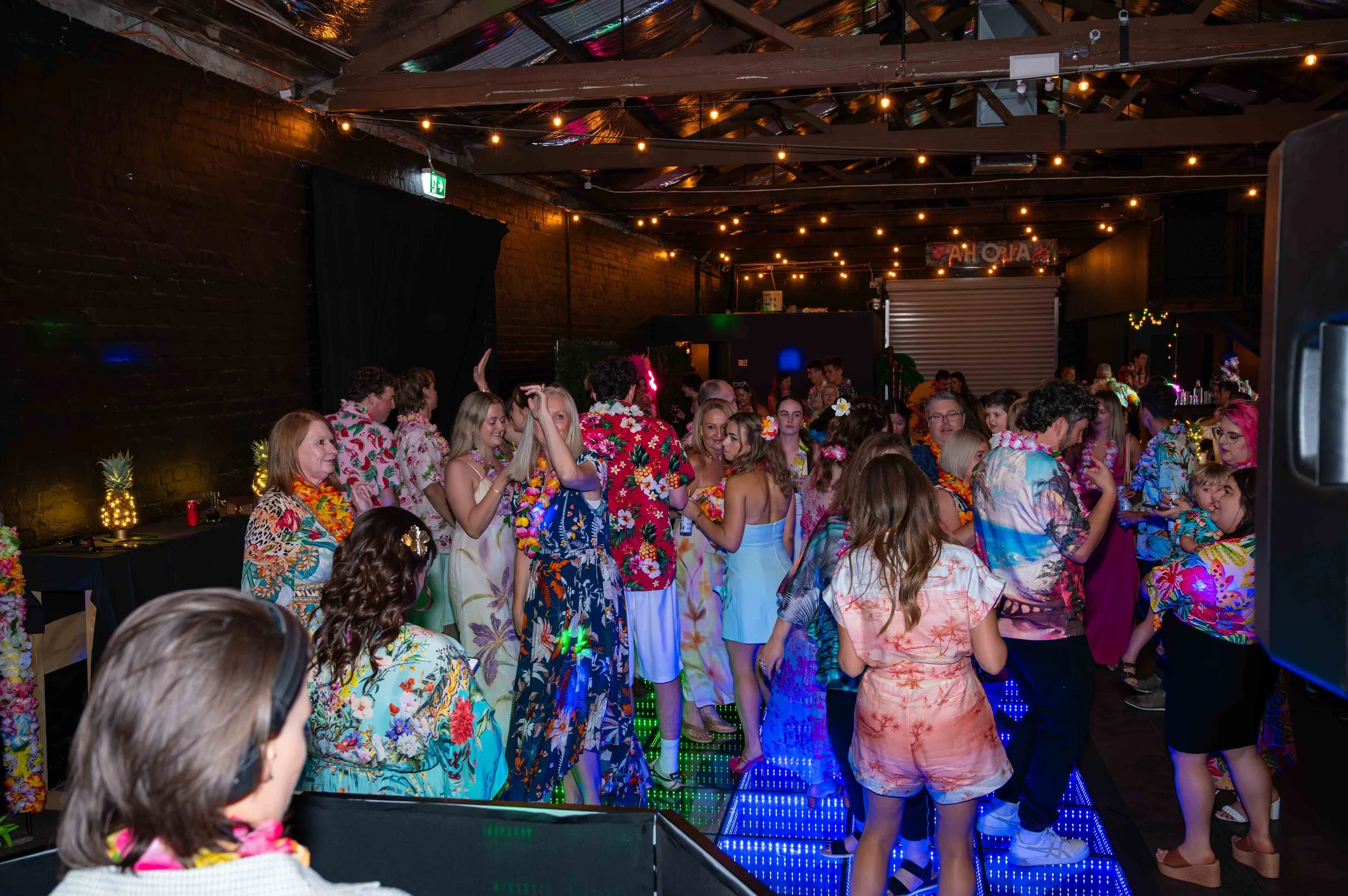People wearing colorful Hawaiian shirts and leis dancing and socializing at an indoor party with string lights and a lit-up dance floor.