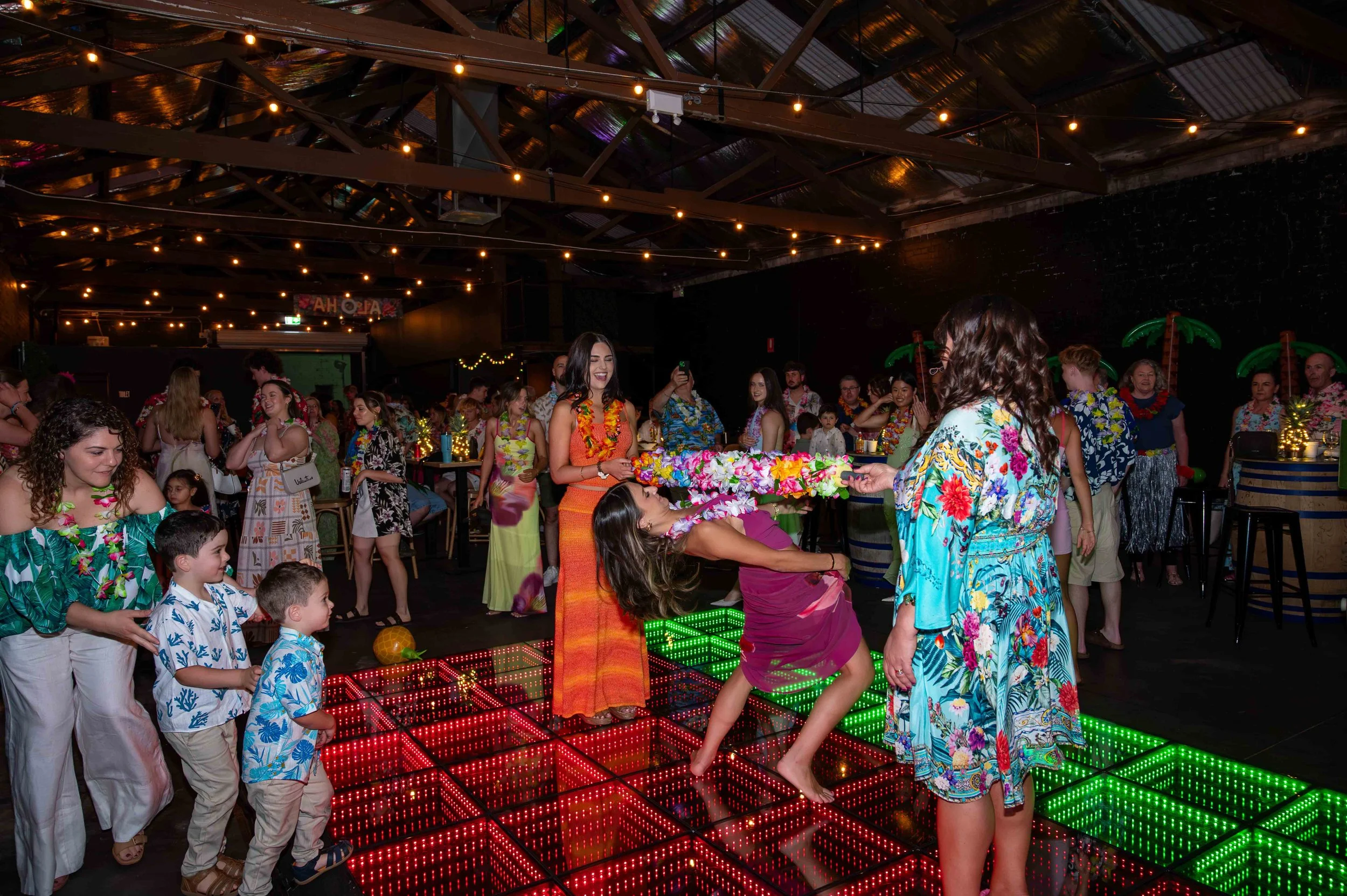 People at a Hawaiian-themed party or event with lights, leis, and tropical decorations, including inflatable palm trees. Some individuals are dancing or engaging in activities on a colorful illuminated dance floor.